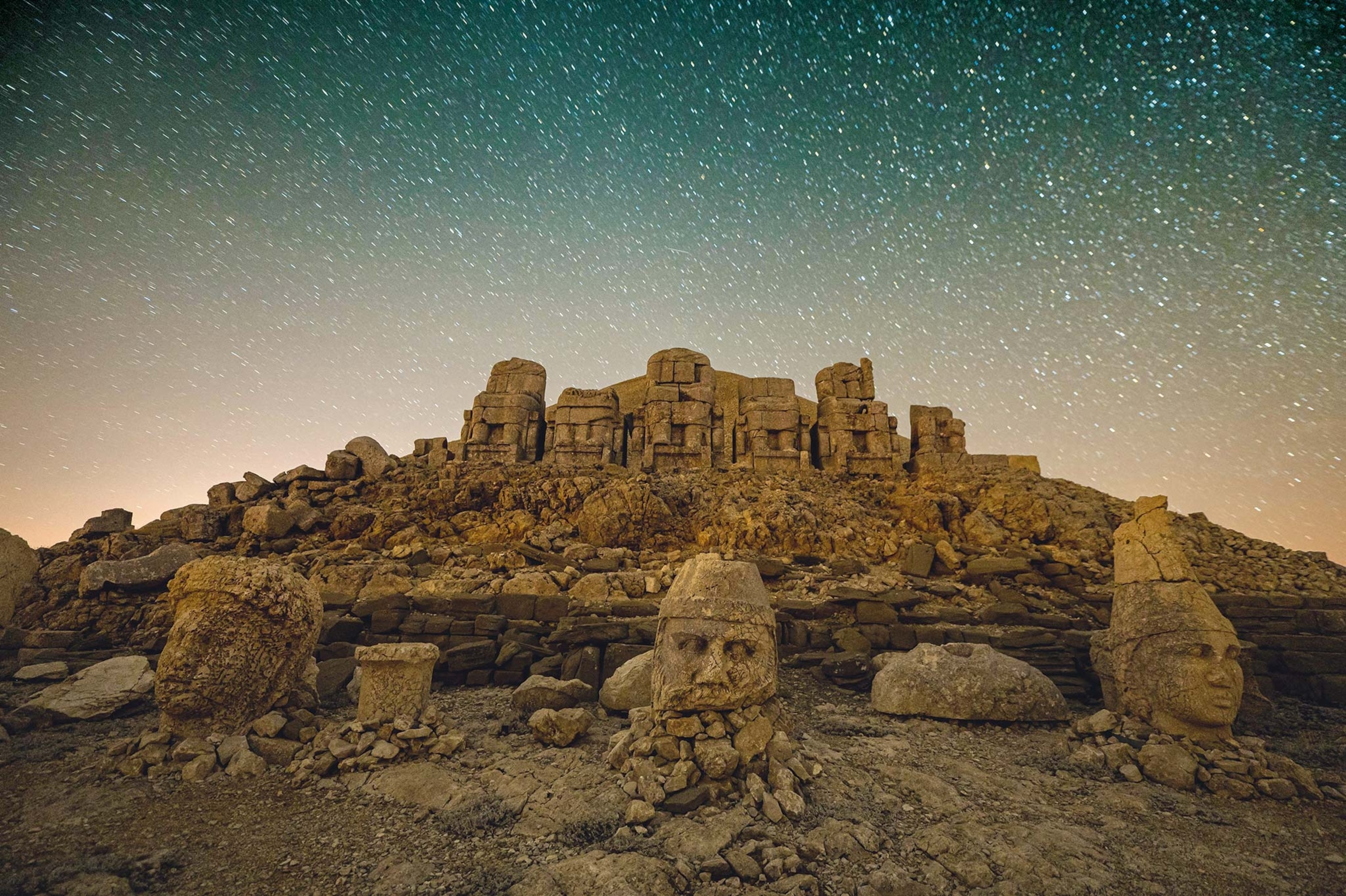 Head statues sit on dirt with the ruins of at temple behind them and the night sky above