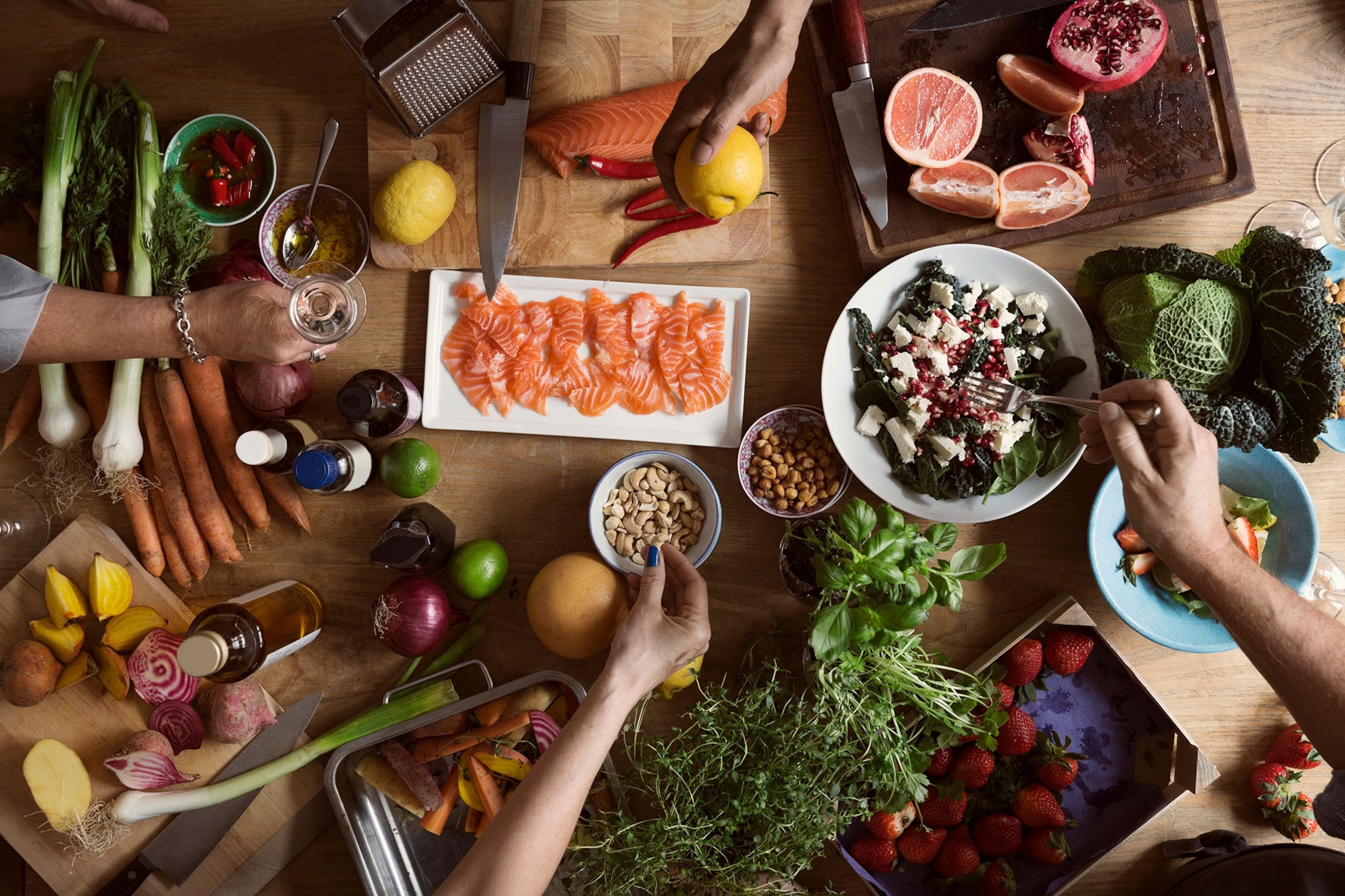 Ariel image of a dinner table with Mediterranean style food. Four hands can be seen, one is holding a glass, one is holding a lemon, one is reaching for a bowl of cashews, and the other is sticking a fork into a salad with beets and cheese.
