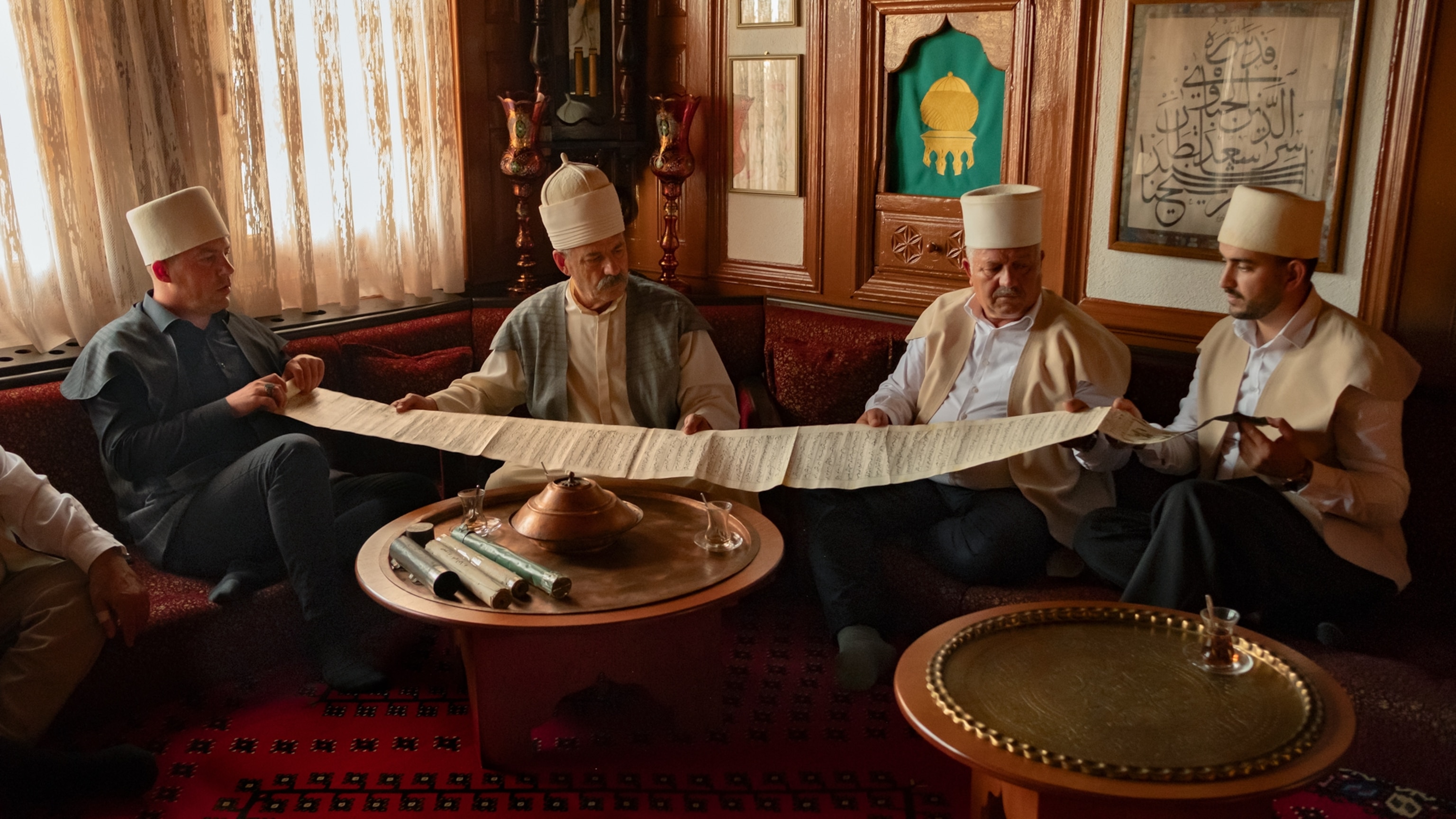 Four men sit left to right and gently hold open a rare scroll as they inspect the writings.