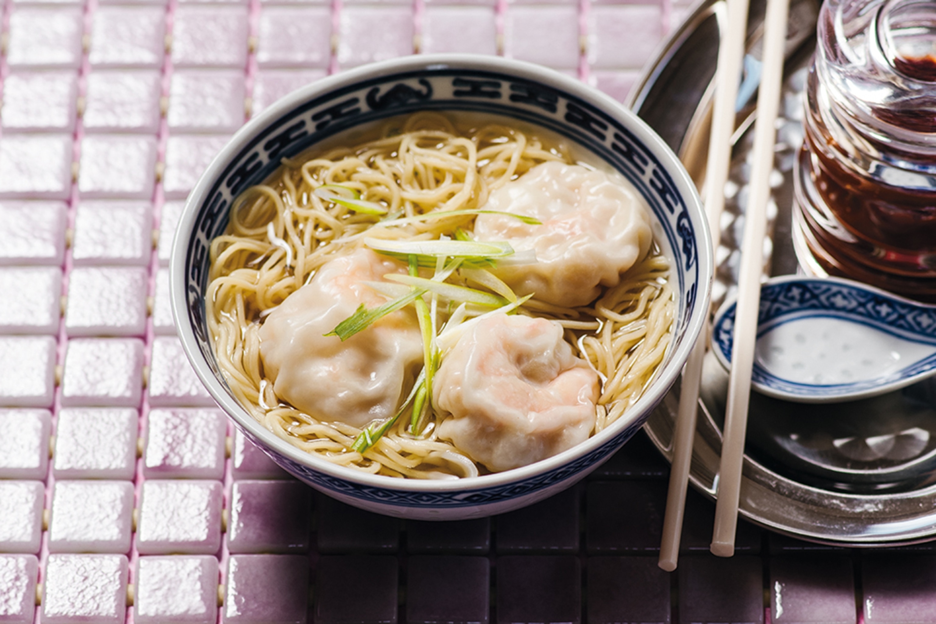 Noodle soup in a bowl with dumplings with chopsticks and a jar of chillis alongside