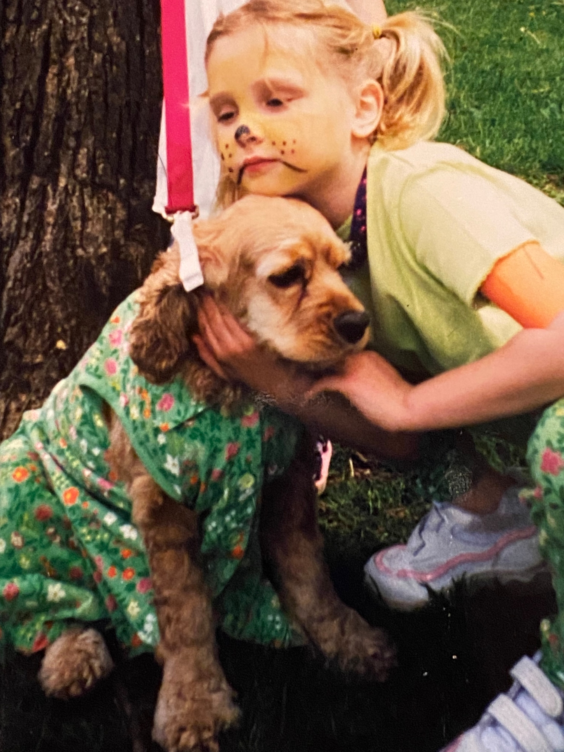 On the grass next to a tree trunk, a young white blonde girl, whose face is made up to look like a dog, crouches next to a dog wearing a green outfit with a floral pattern.