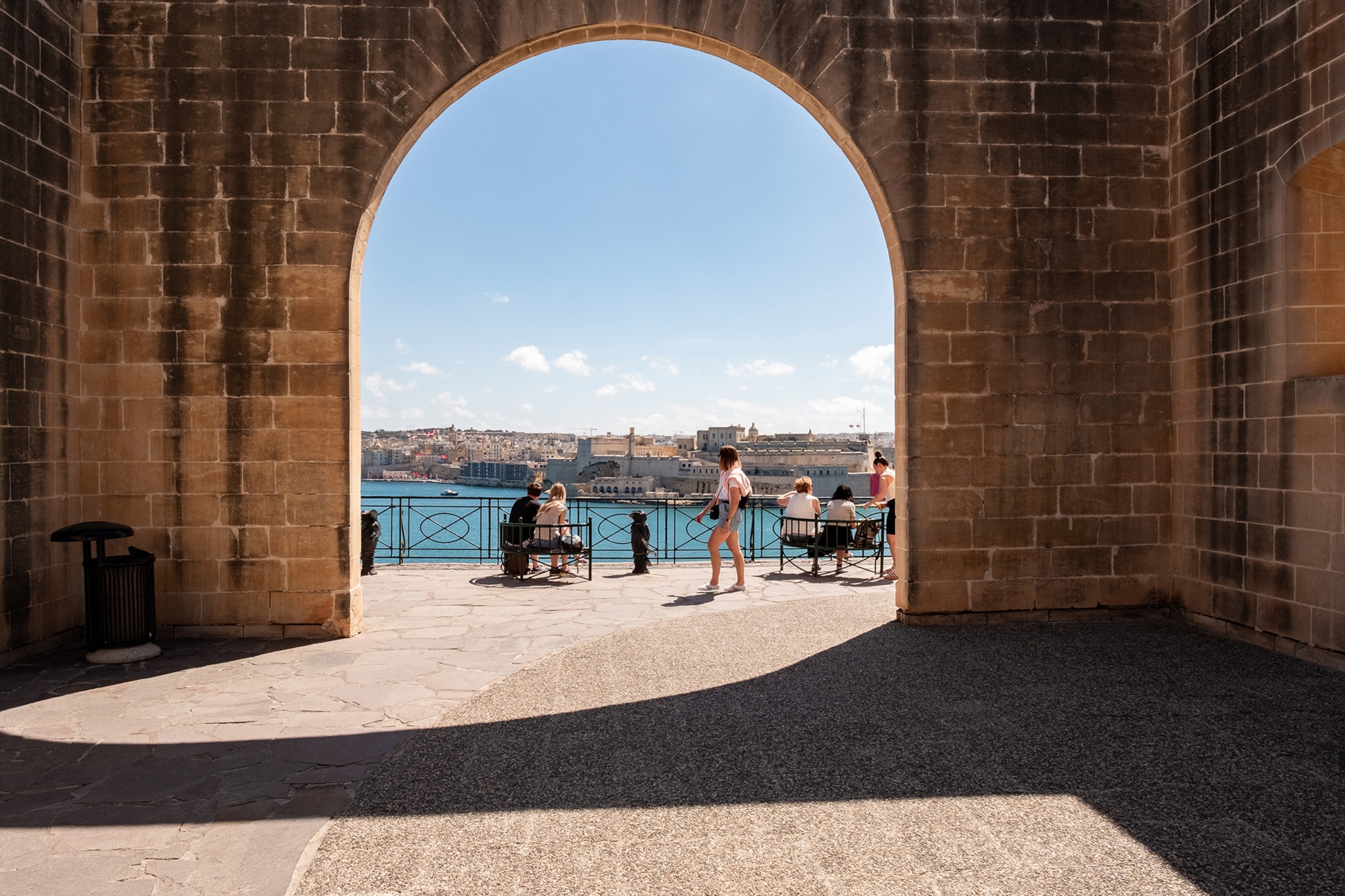 A view of Valletta's harbour from an old limestone archway. People sit on beaches by the water, whilst some walk past, not without admiring the view.