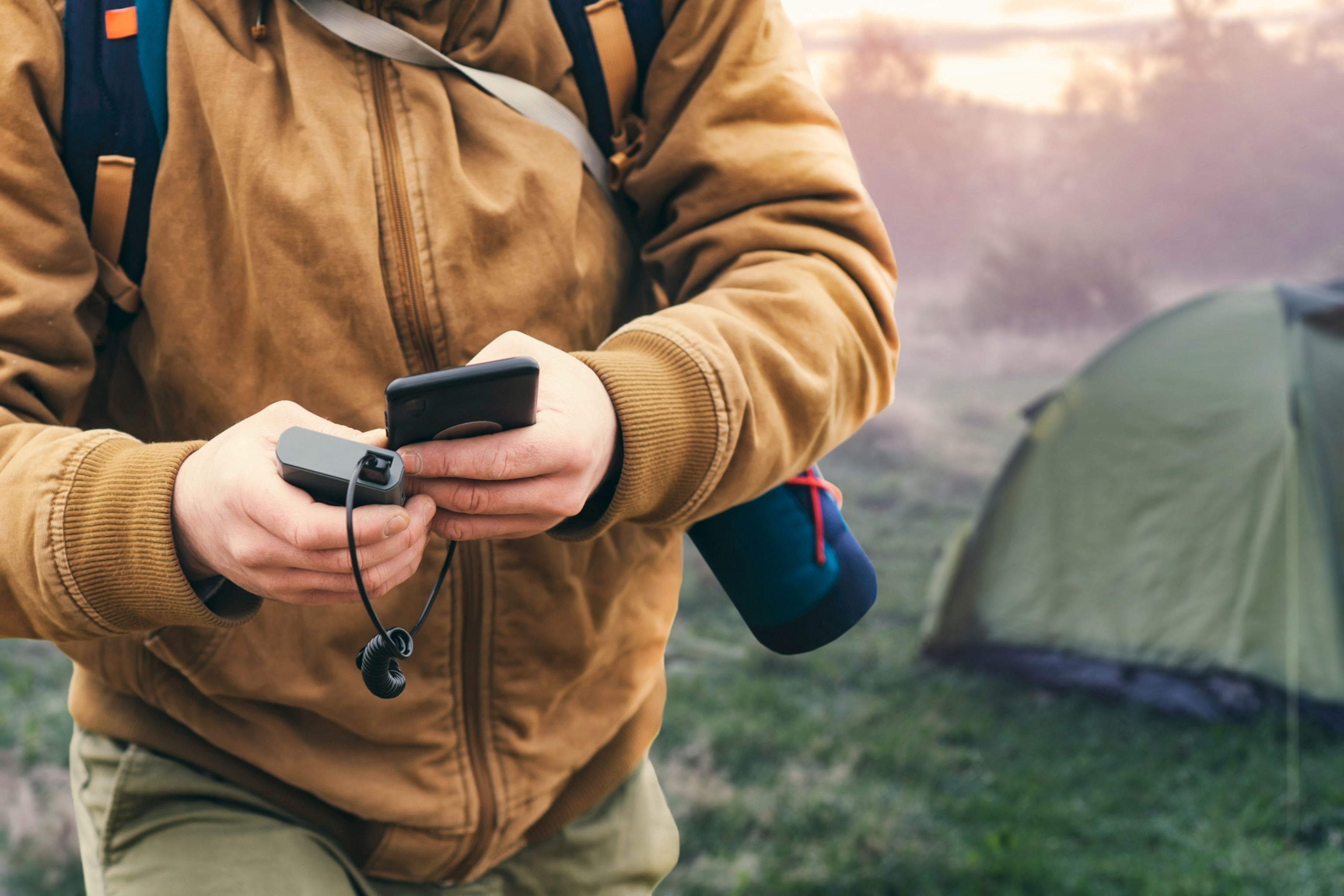 A close view of a man holding a smartphone in his hands as he charges it with a power bank, against the backdrop of a tourist tent in nature