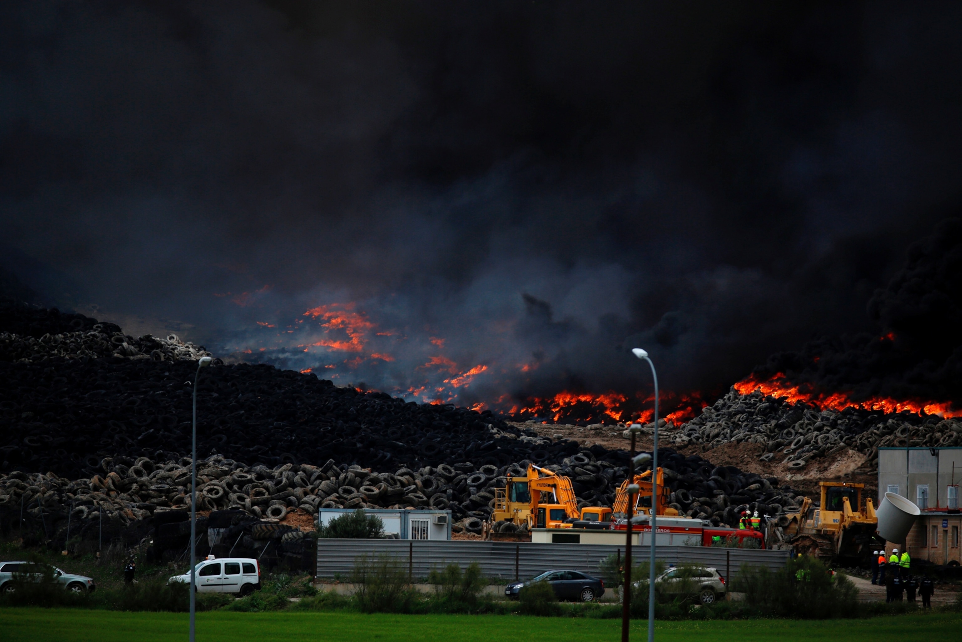 Beneath a sky filled with black smoke, fire rages amongst stacks of rubber tires.