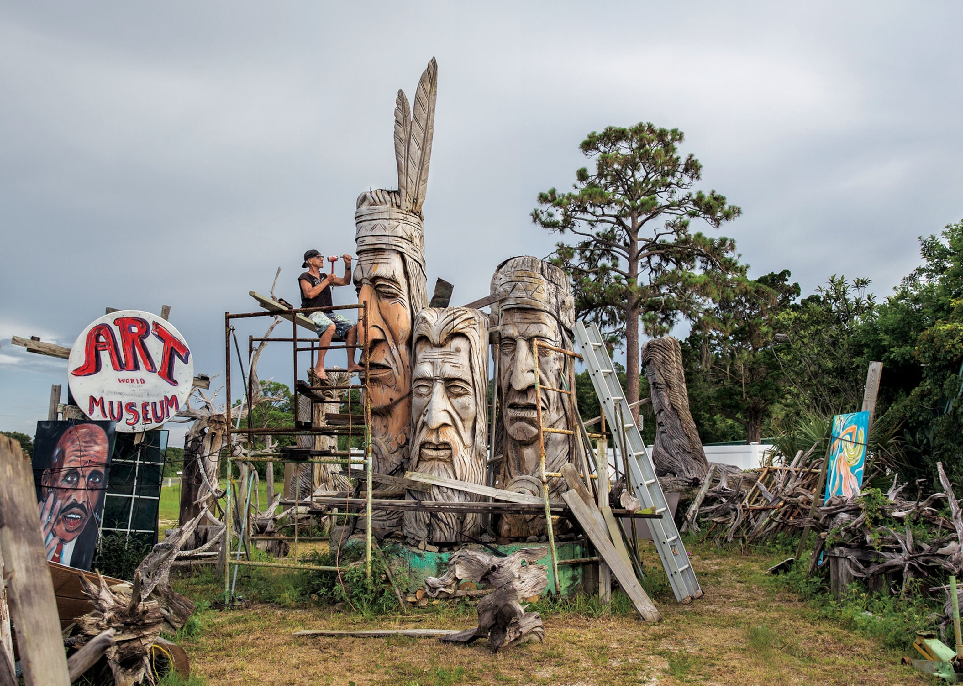 a man sculpting in a yard filled with other large face sculptures