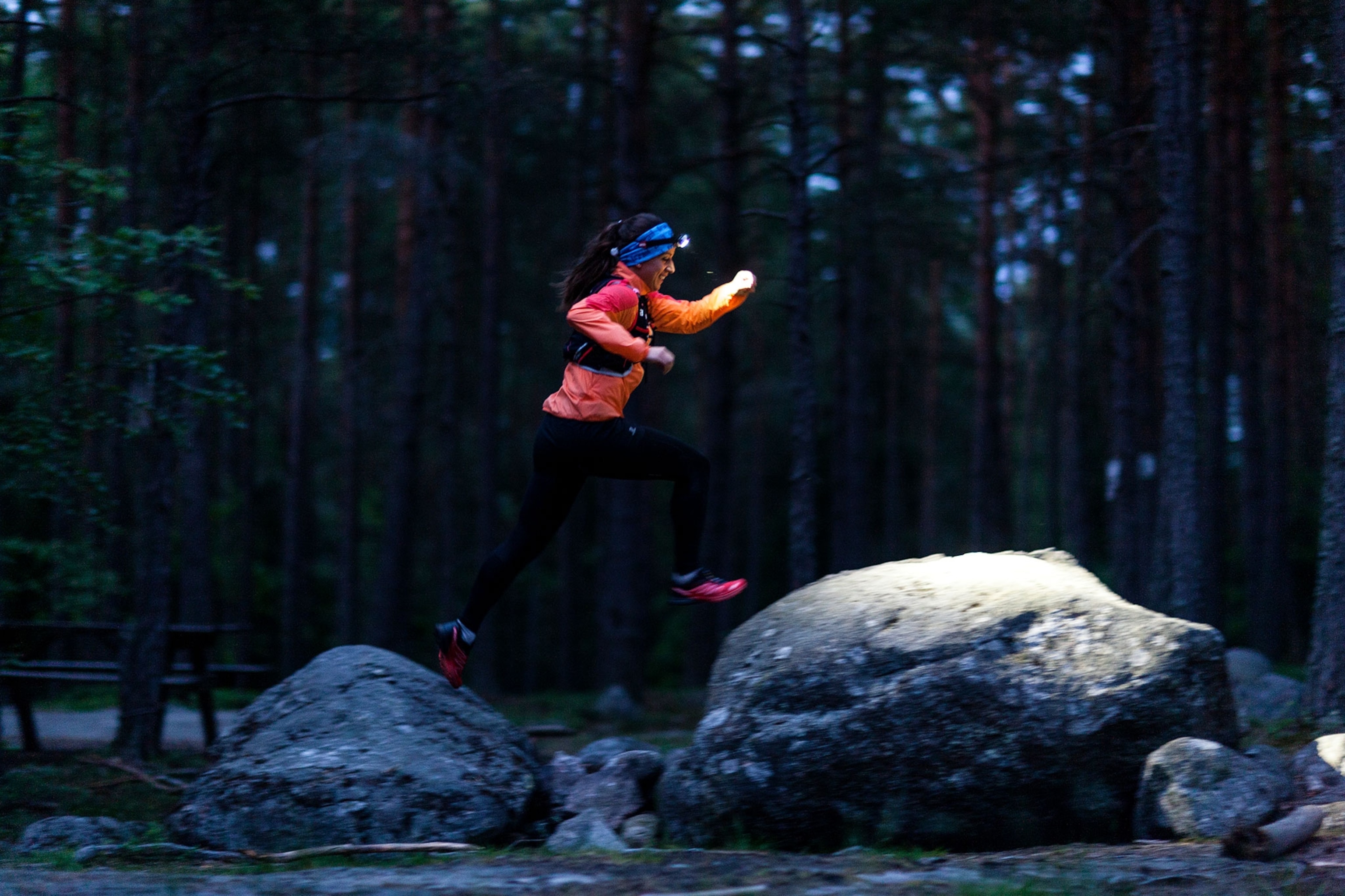 A woman jumps over large rocks at nighttime while wearing a headlamp, lighting up the rocks below.