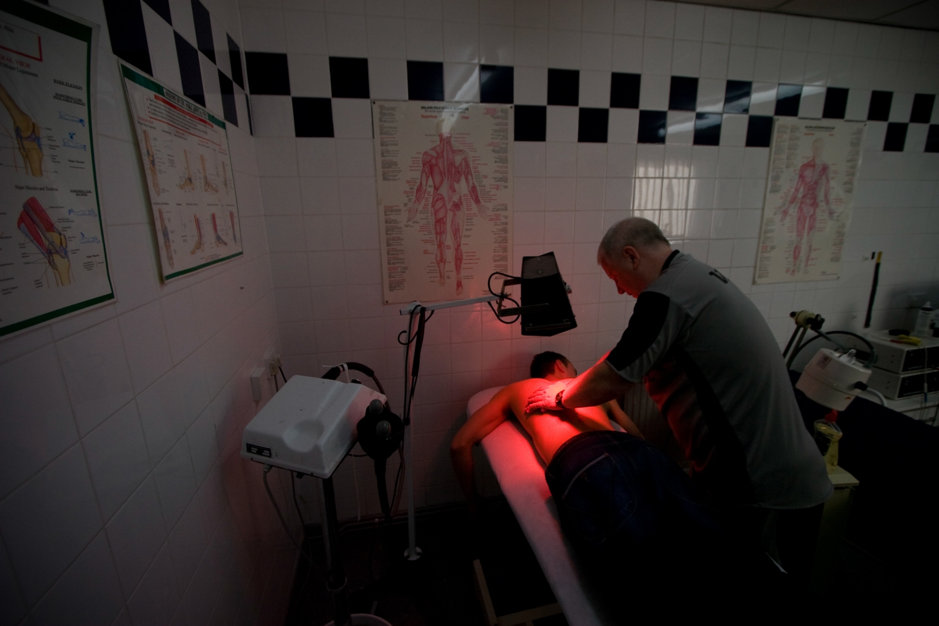 A red light shining down on the back of a young athlete laying face down on an examination table as a physiotherapist massages his back.
