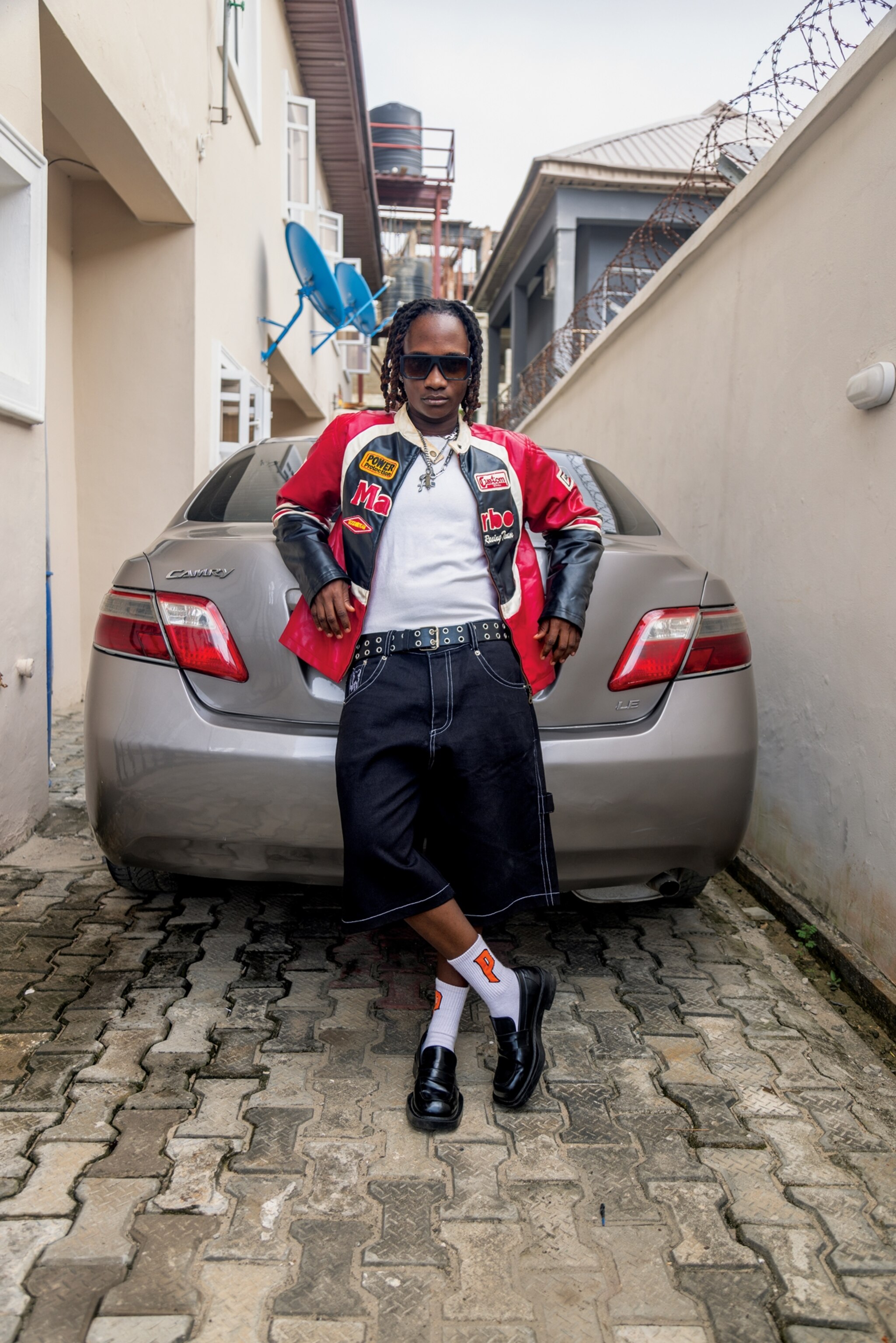 Jordyn is leaning against the trunk of a bronze Toyota Camry trunk wearing a black and red leather Marlboro jacket, sunglasses, long jean shorts, tall socks, and loafers.
