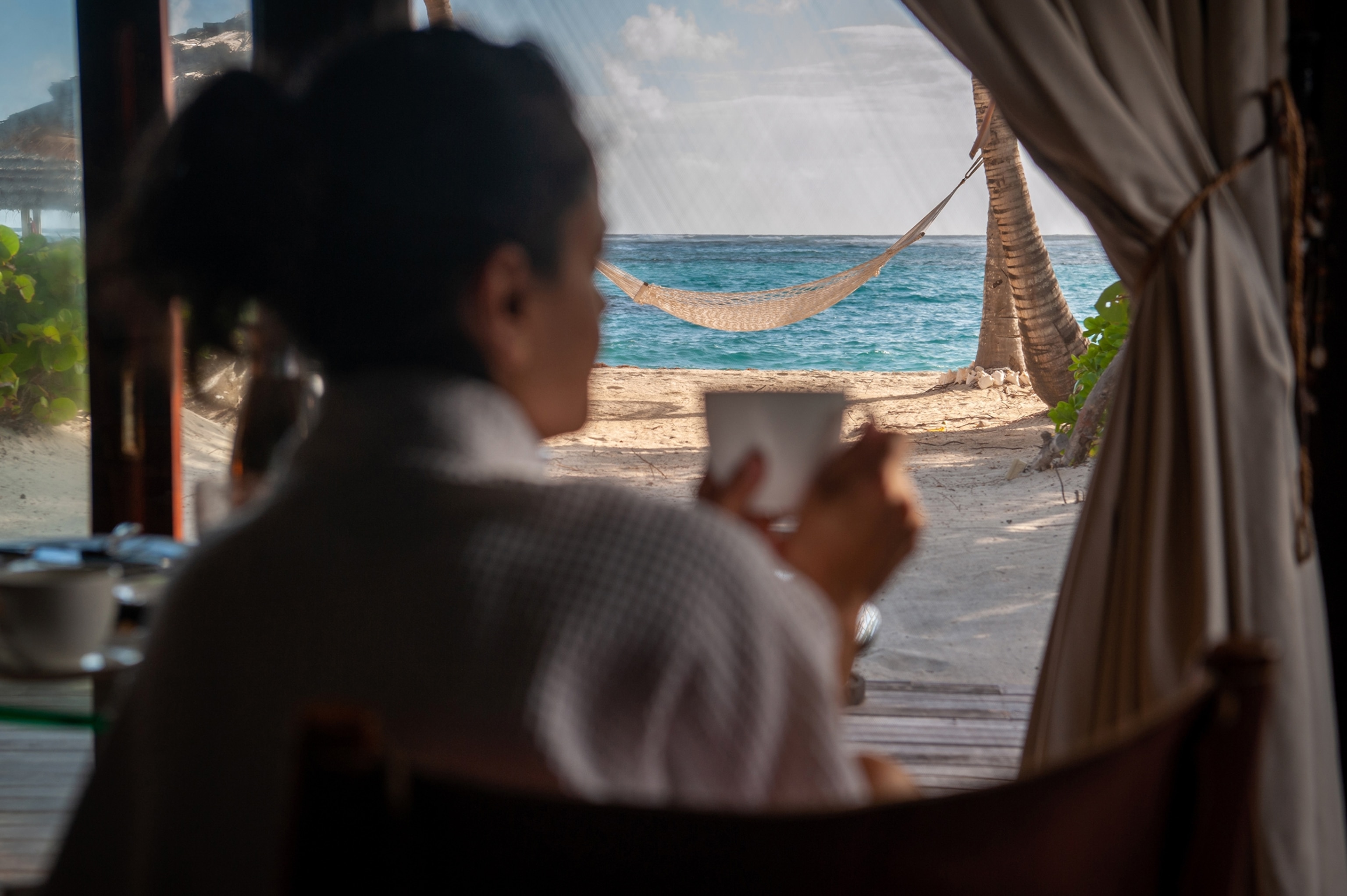 In the foreground, a blurred figure of a woman wears a white robe and holds a white coffee cup while looking at a beachfront view.