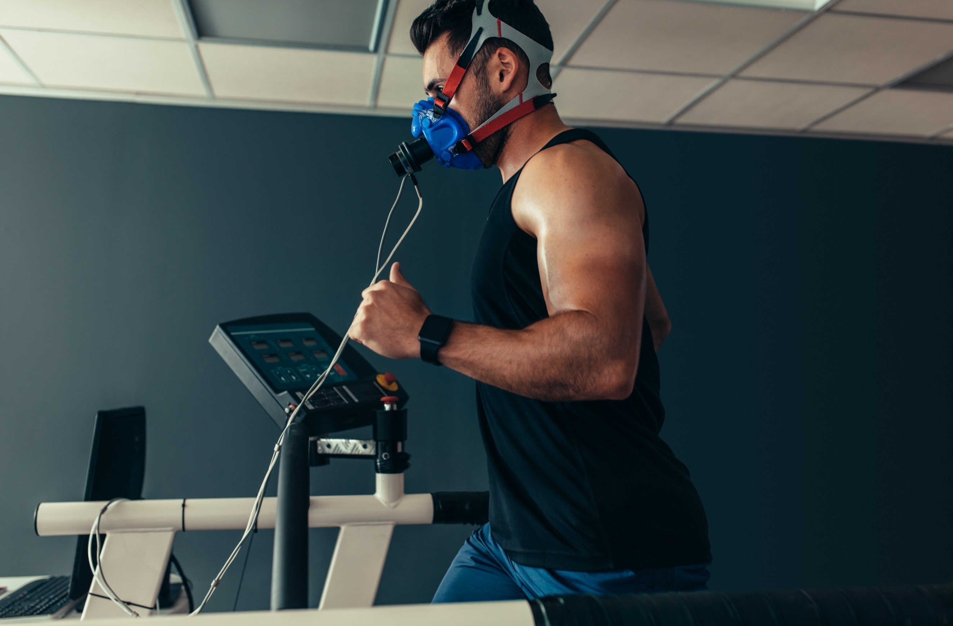 A male athlete running on a treadmill in a sports science lab. He has a mask strapped over his nose and mouth to measure his oxygen consumption during exercise.