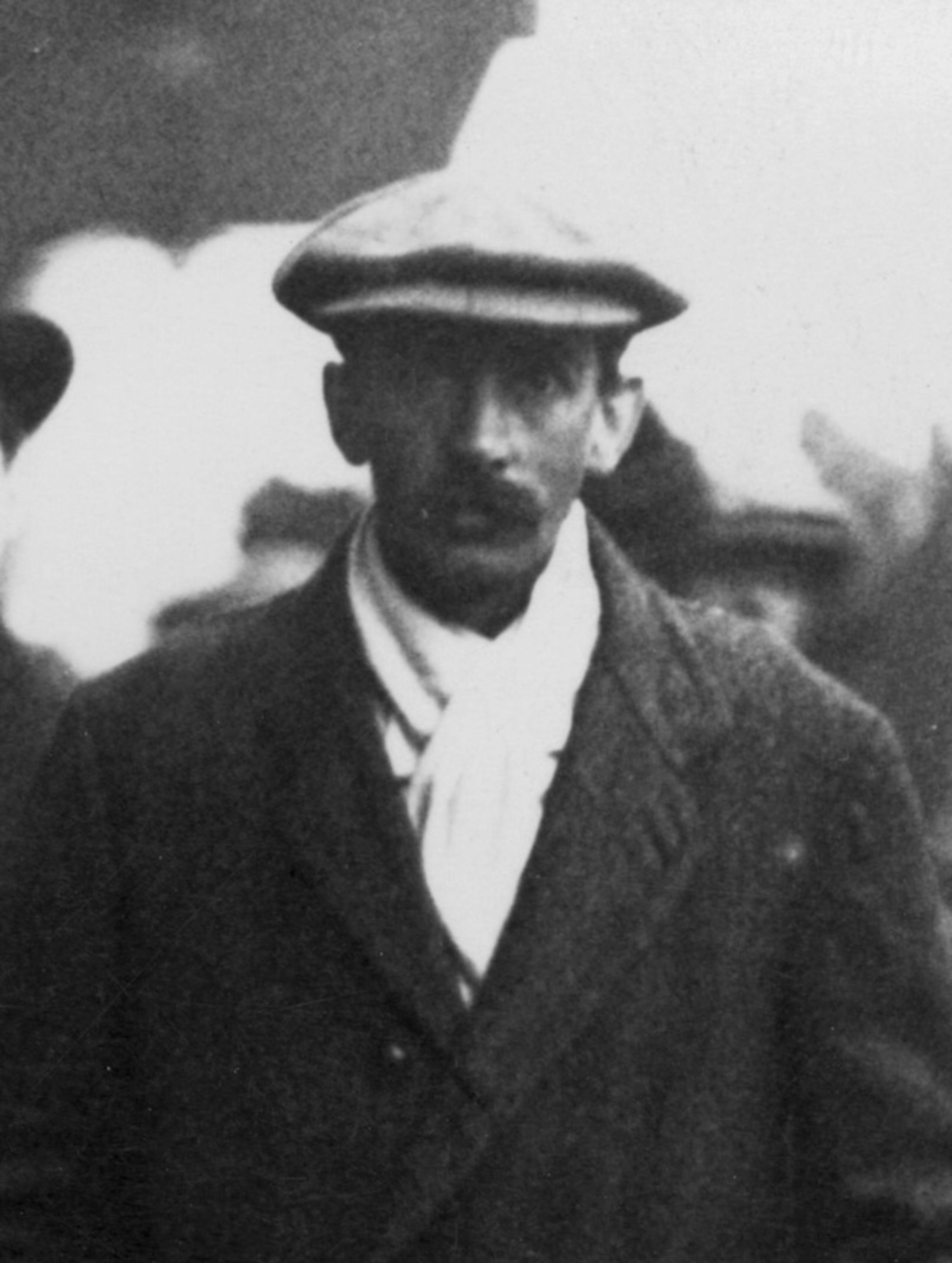Head-and-shoulders portrait of a mustachioed white man in a flat cap and suit jacket in 1913.