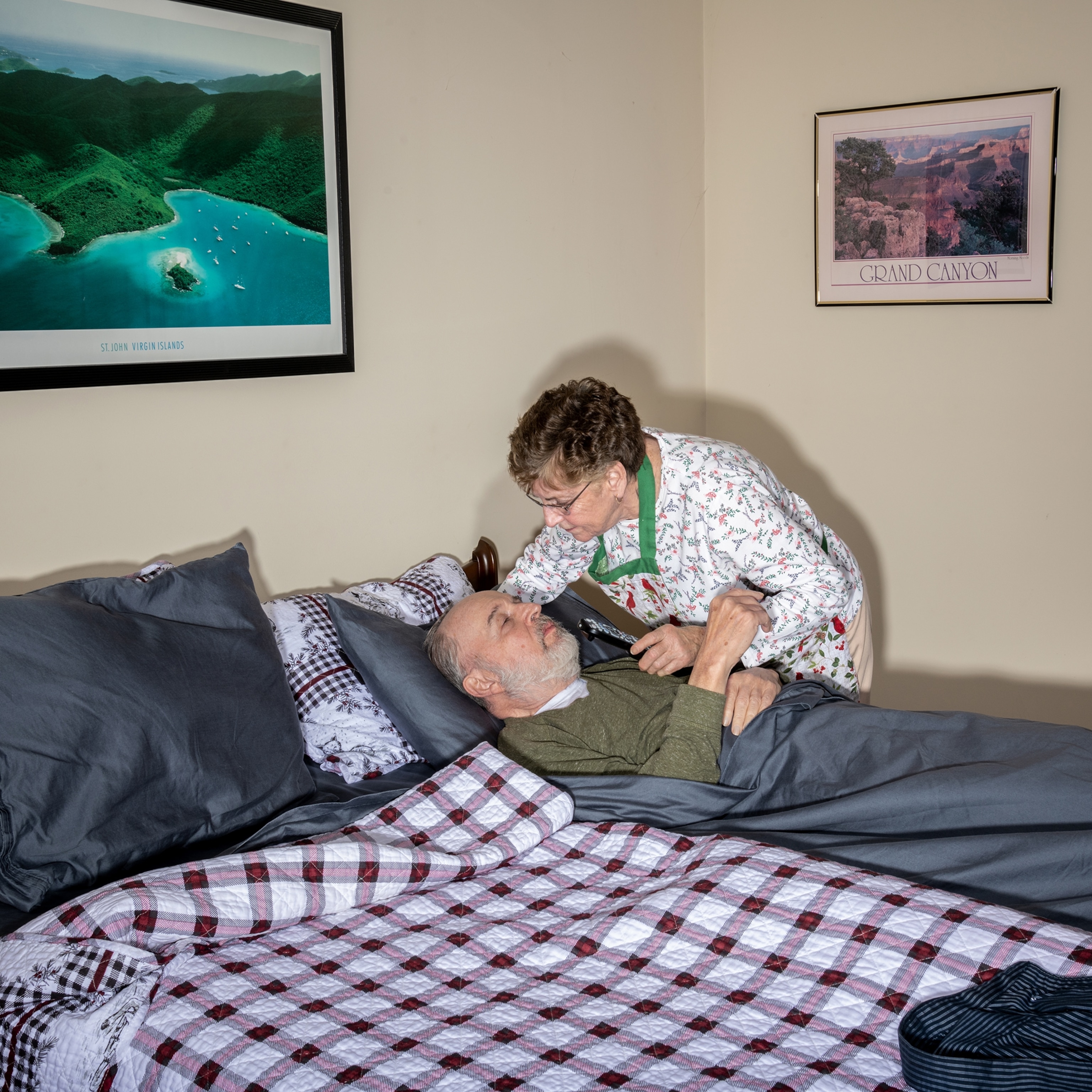A man lies in bed covered with a dusty blue sheet and a red plaid quilt as his wife leans close by his side.