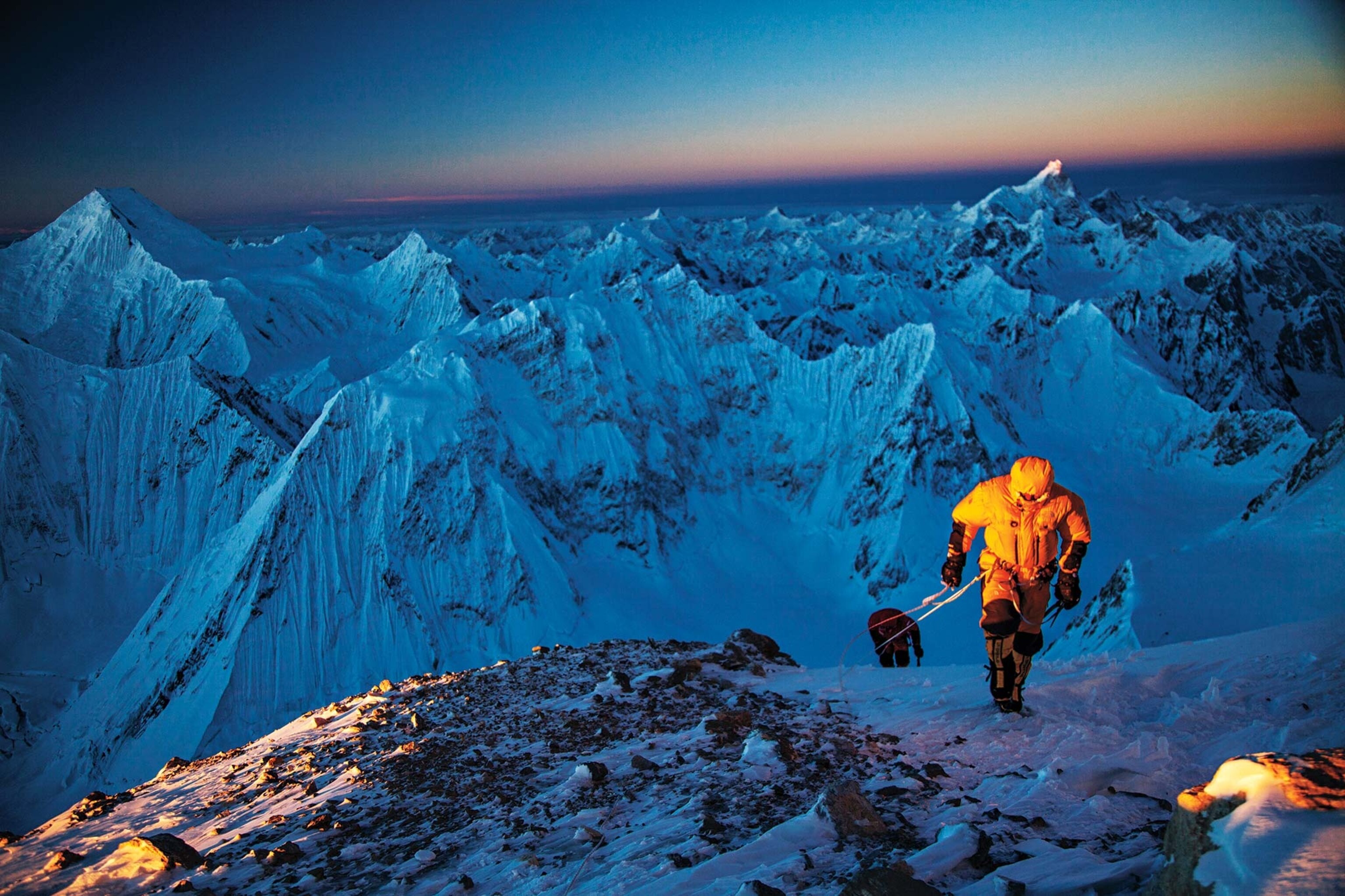 Two mountaineers, one in yellow, near the top of a snow-capped peak