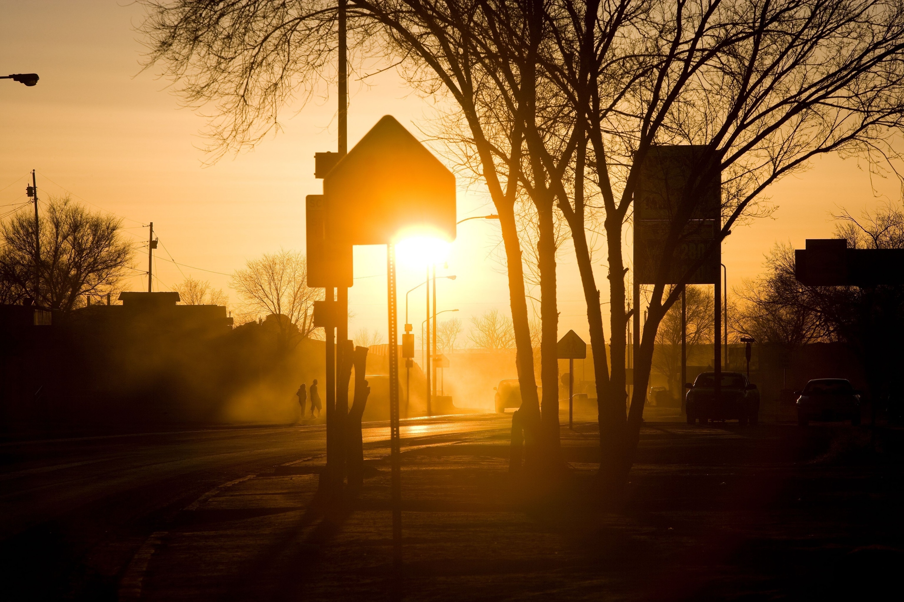 The bright orange light of a sun setting silhouettes road signs and trees on a neighborhood street. In the distance are two pedestrians and a car driving on the road.