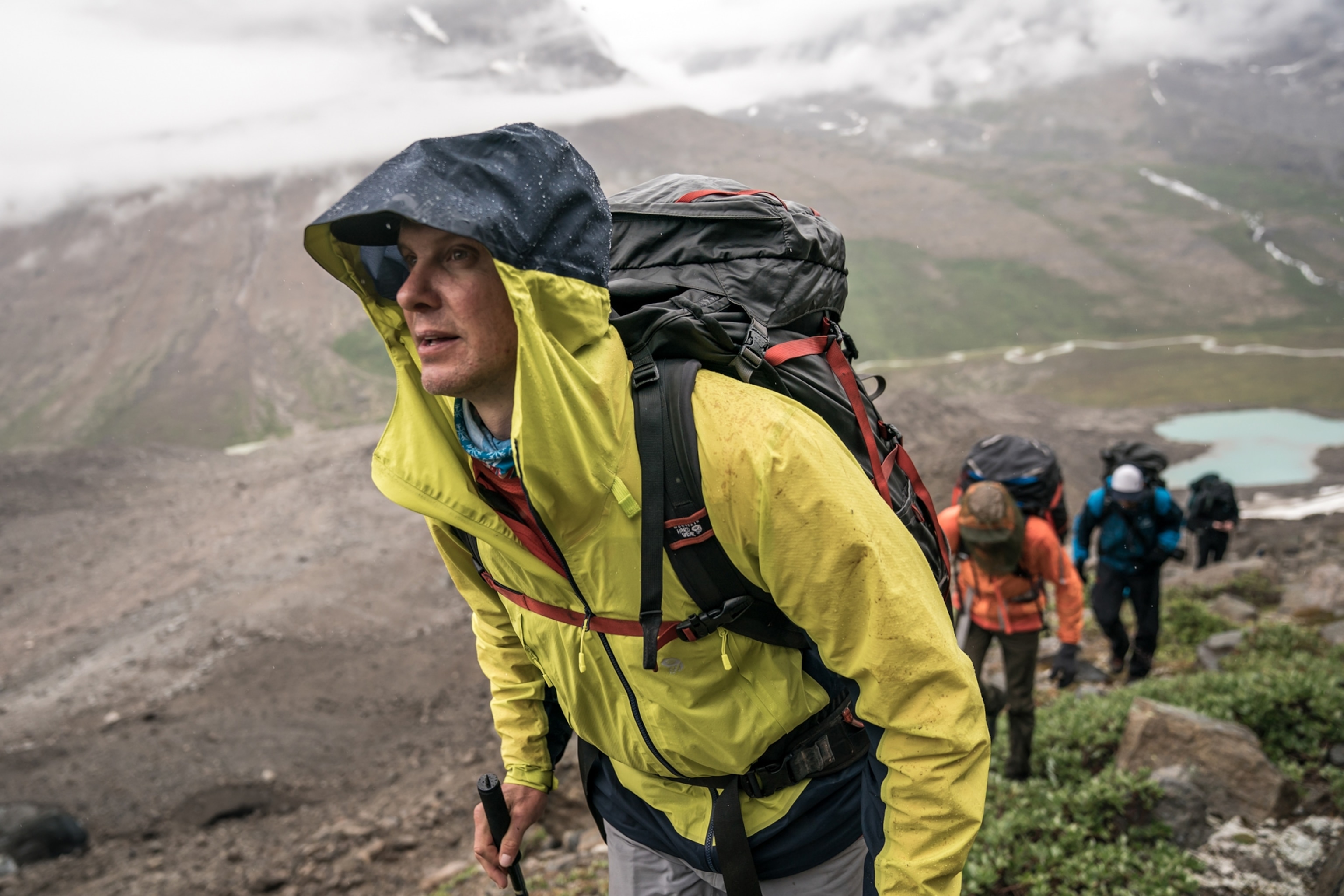 A man in a yellow wind breaker is walking up a mountain with a backpack on. There are others behind him with backpacks. There are mountains in the background with a thick fog around them.