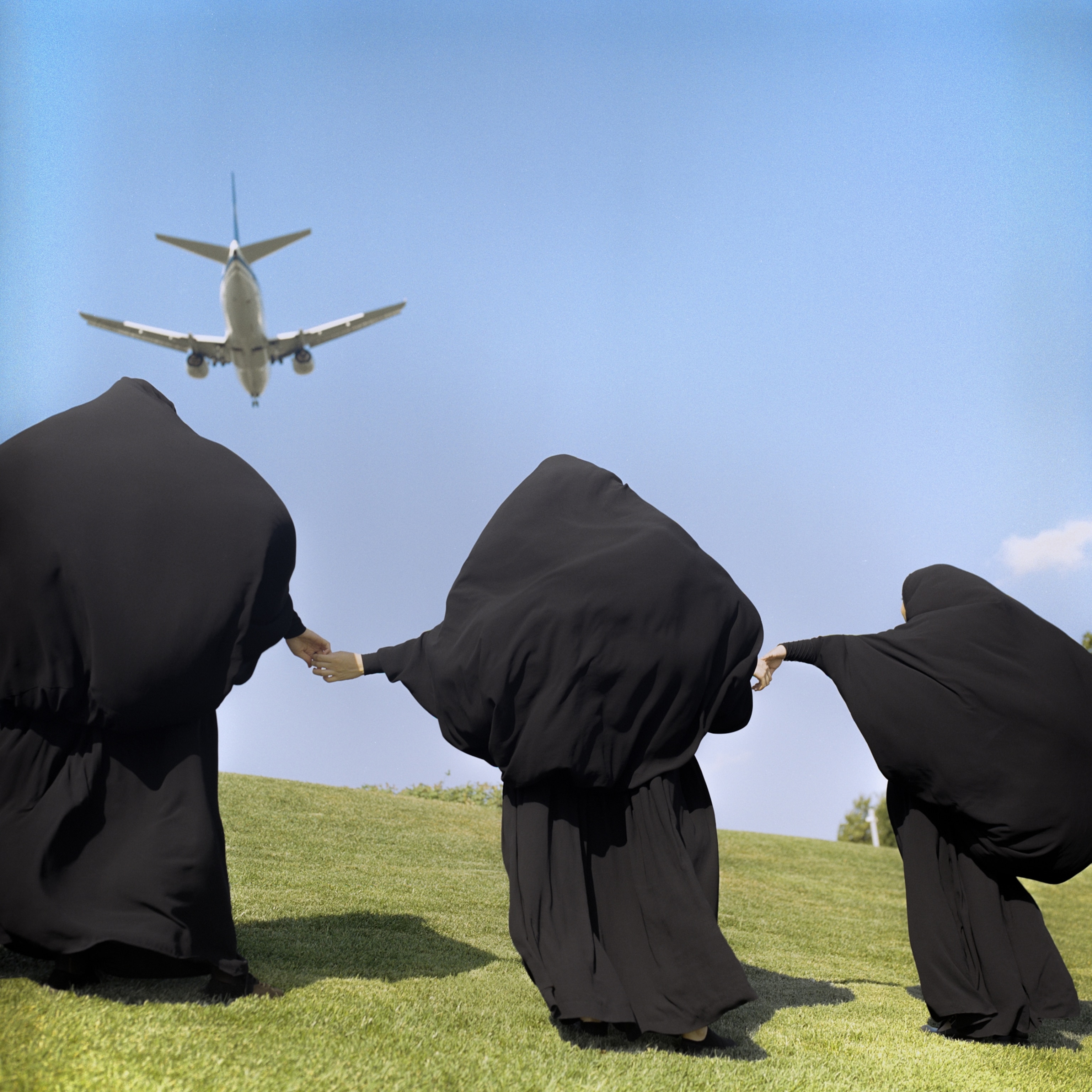 Picture of three woman in all black shown from behind holding hands and looking up at airplane in sky.