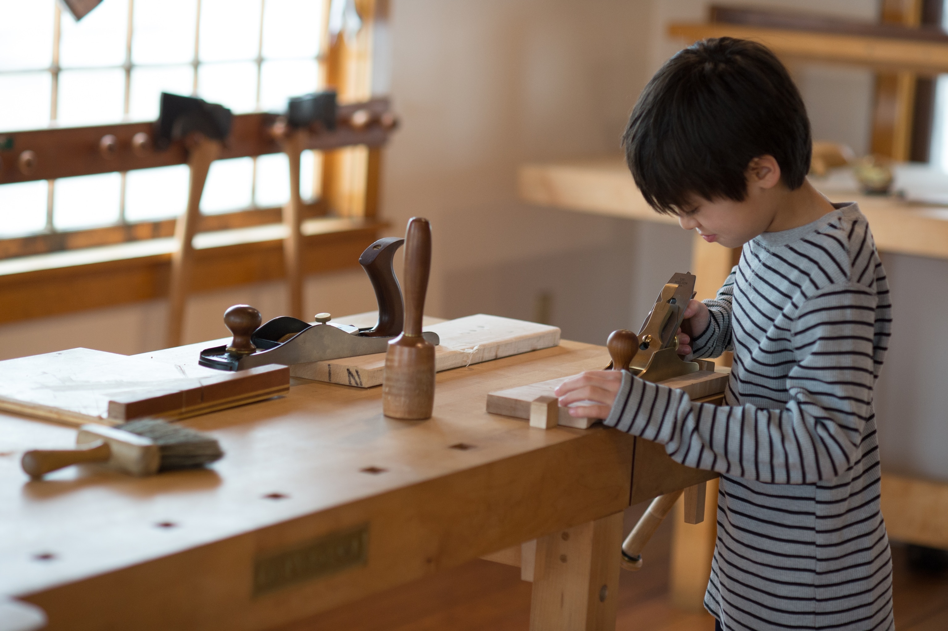 A young boy uses a woodworking tool at a woodworking bench next to the window in a tool factory