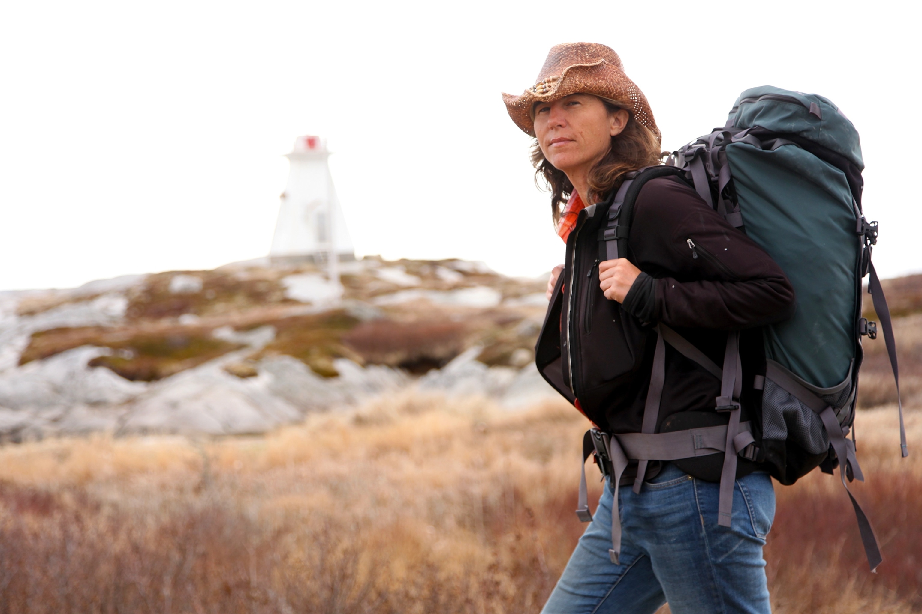A woman posing proudly yet casually in a rocky and arid landscape, holding on to her hiking backpack and wearing a straw cowboy hat.