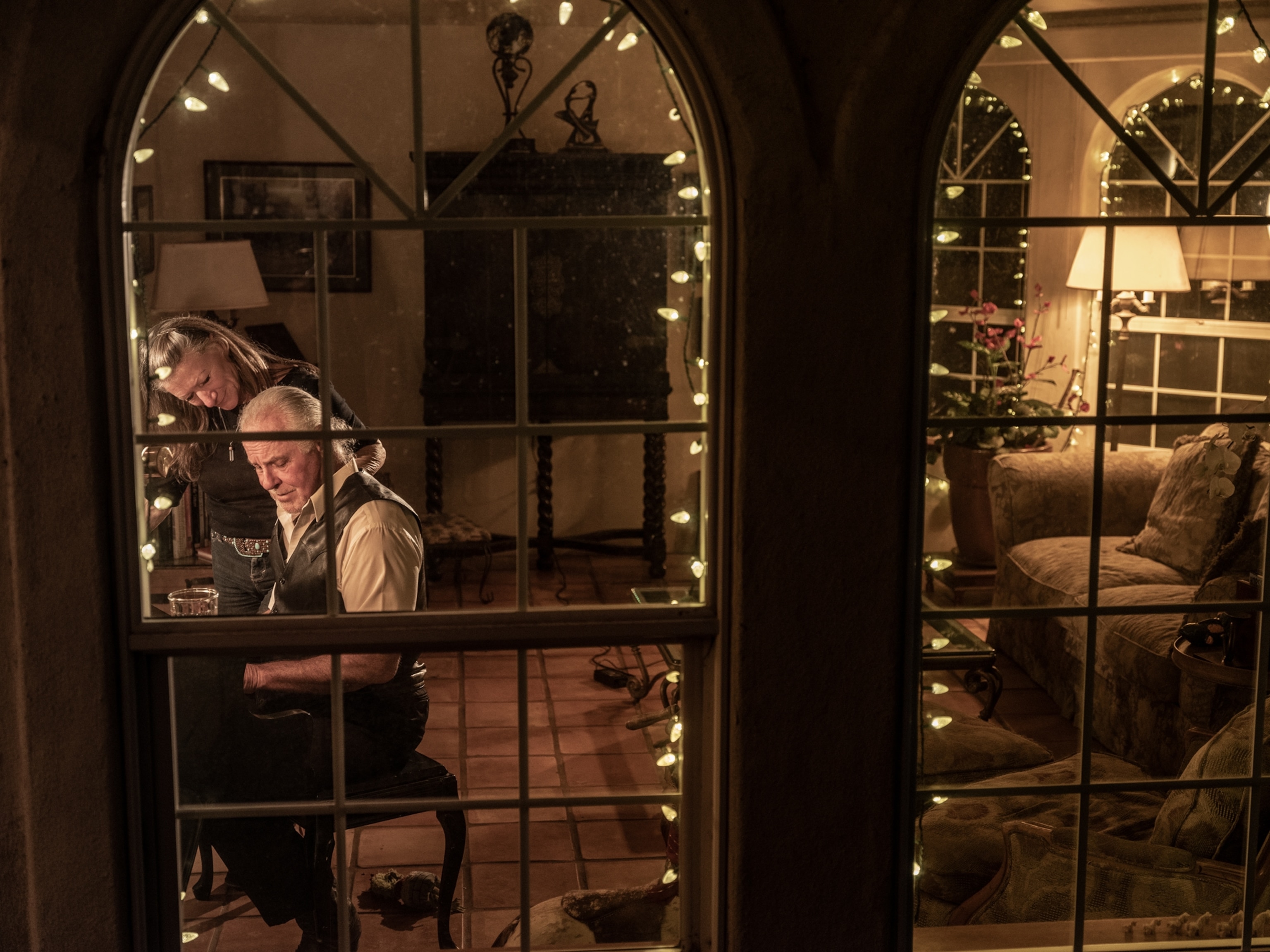 view through home windows of woman listening to man playing piano.