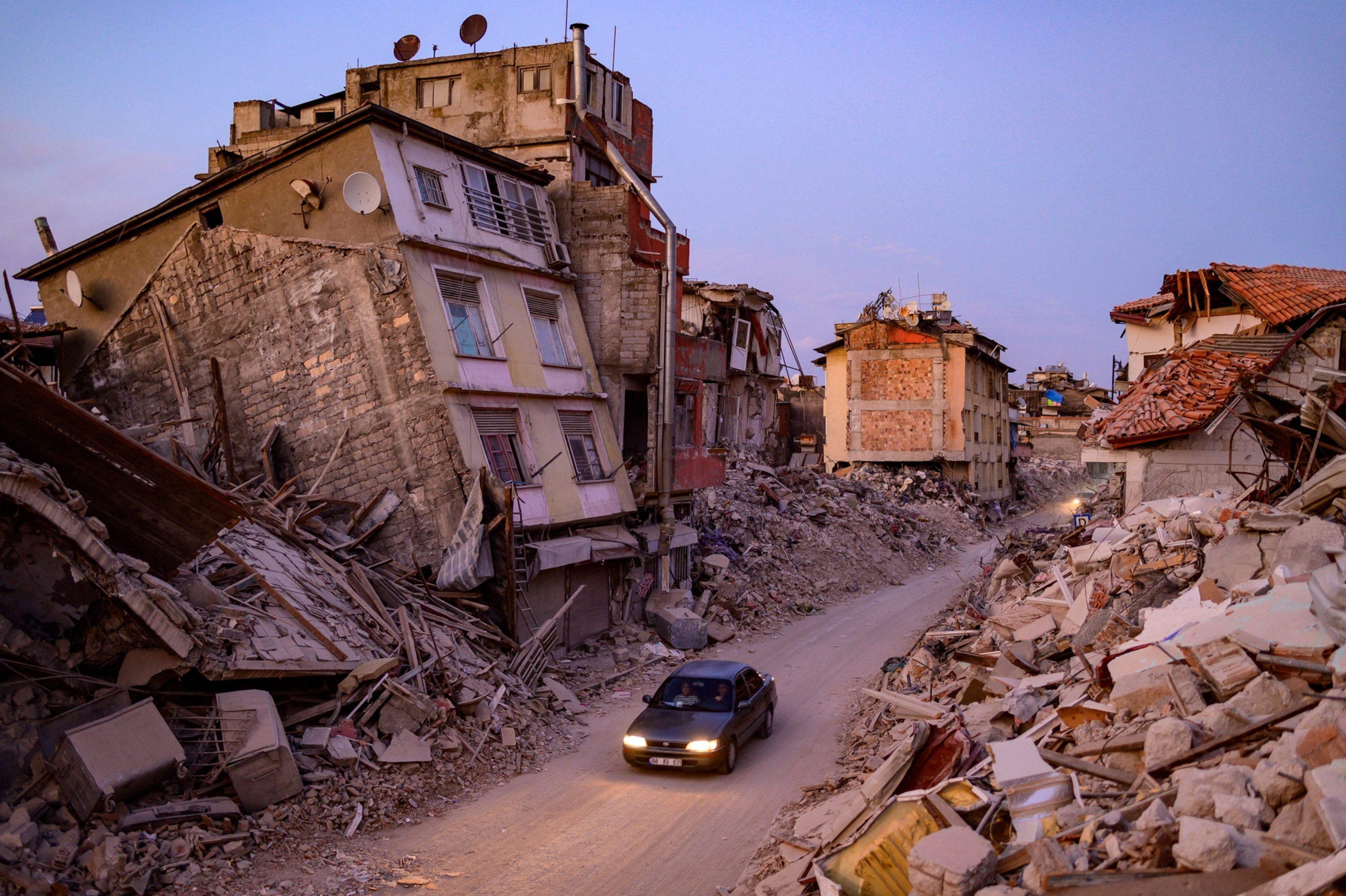 A car drives along a dirt road, which is surrounded by debris from collapsed buildings.