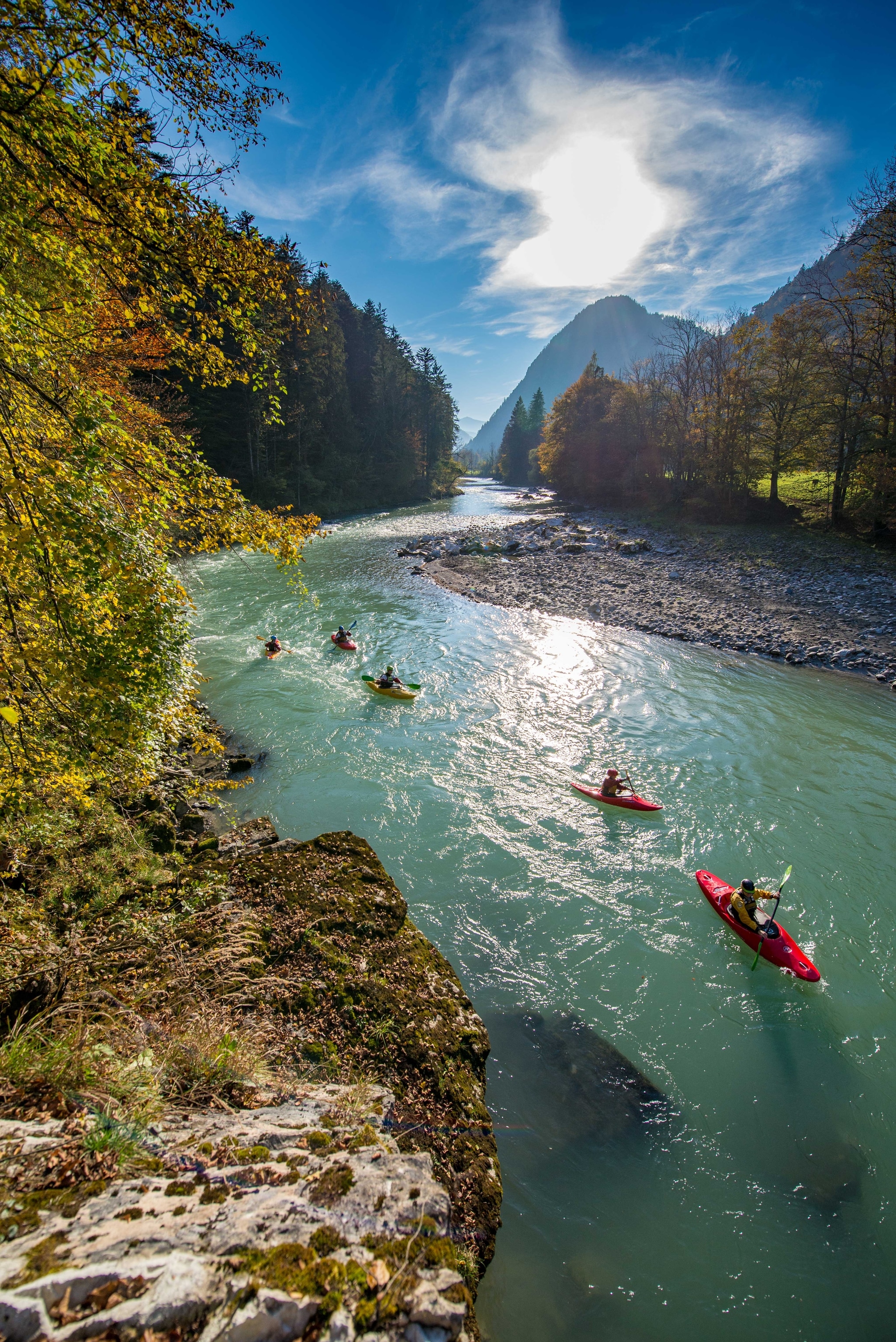 Five kayaks on a blue river, with a pebble beach to one side and mountains in the background.