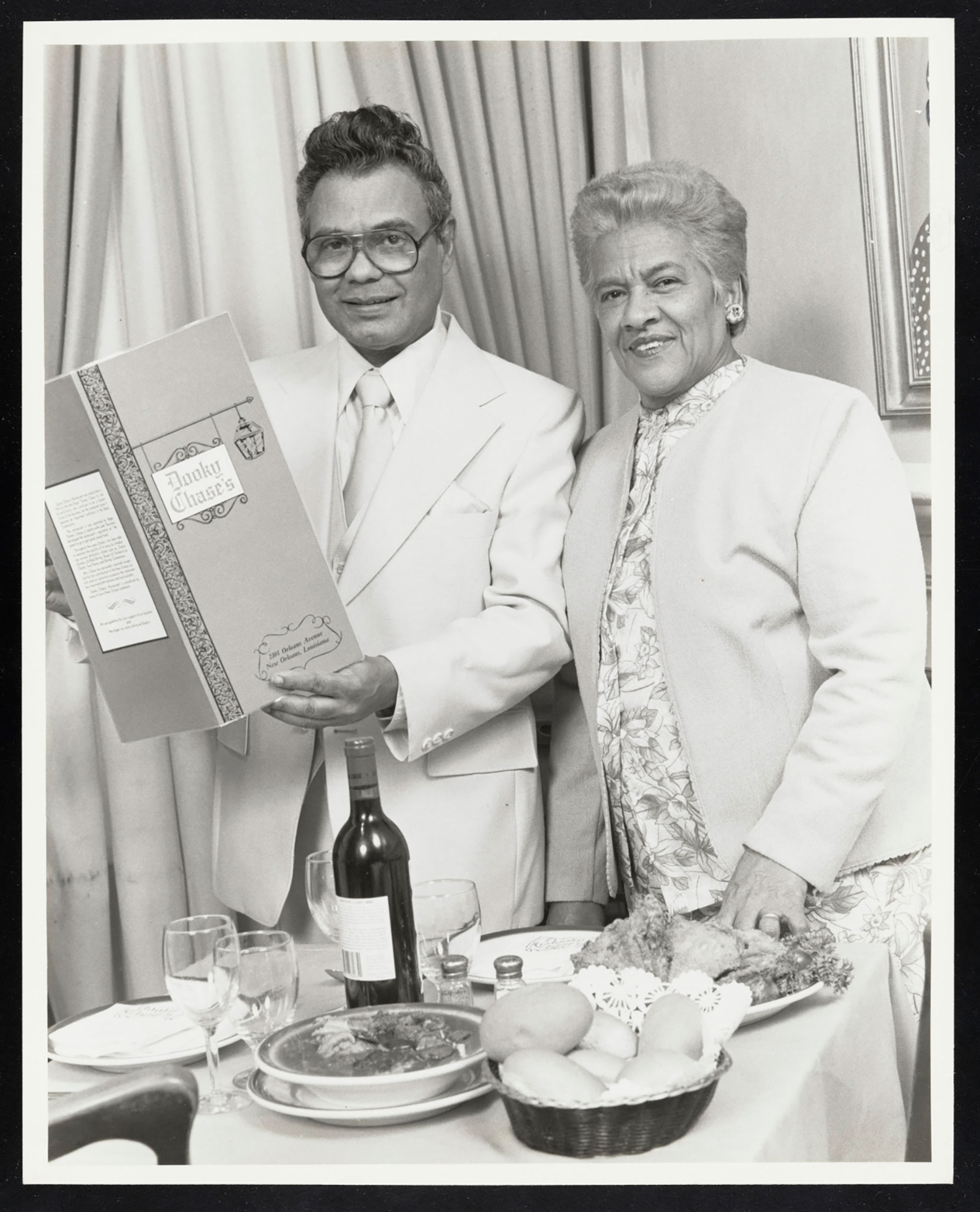 Black and white photo of a couple, dressed in 70s era business attire in front of a table of food at a restaurant. The man is holding up a menu.