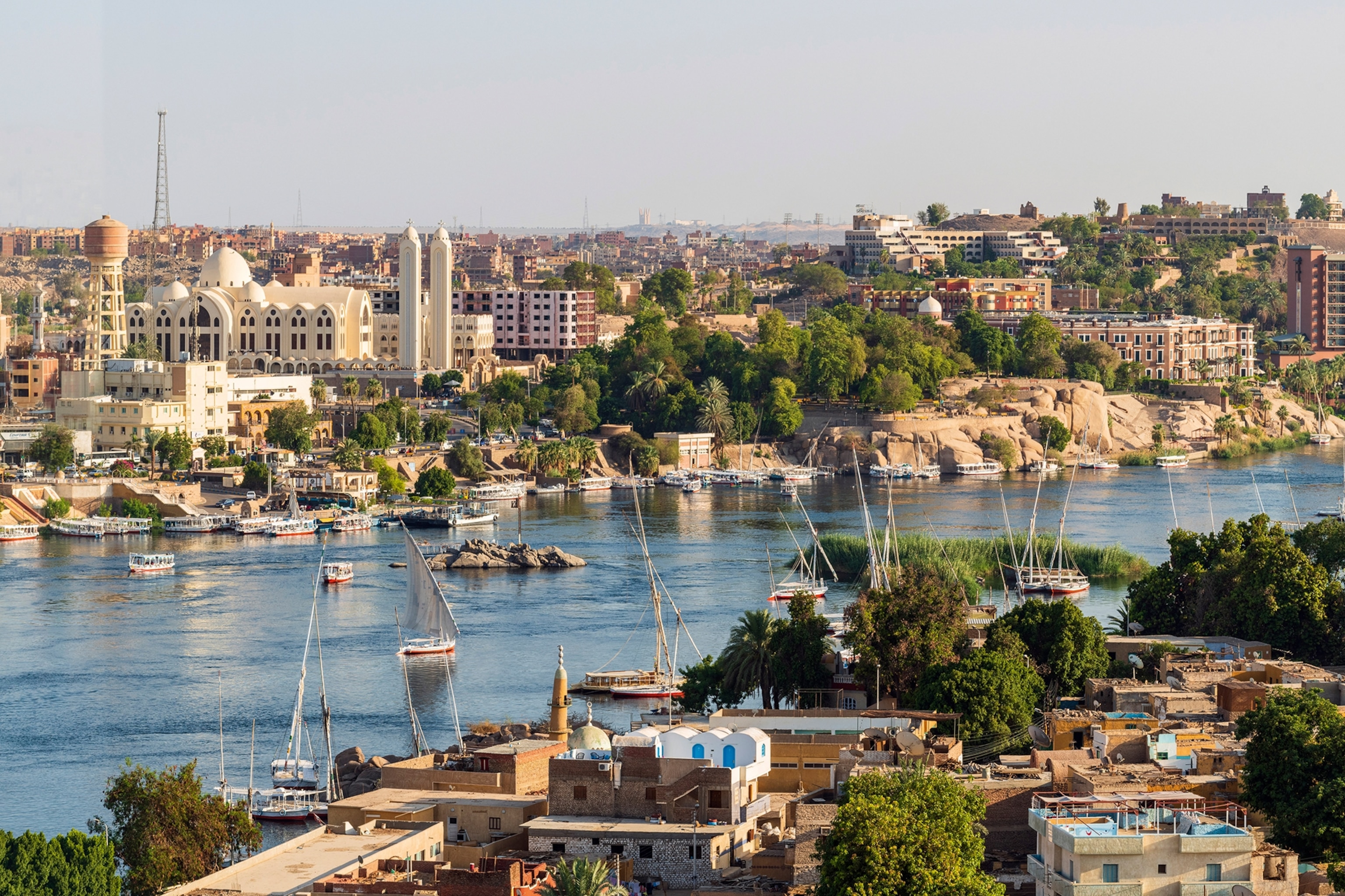 Felucca boats sailing on the River Nile, at Aswan. Trees are dotted among the buildings in the background.