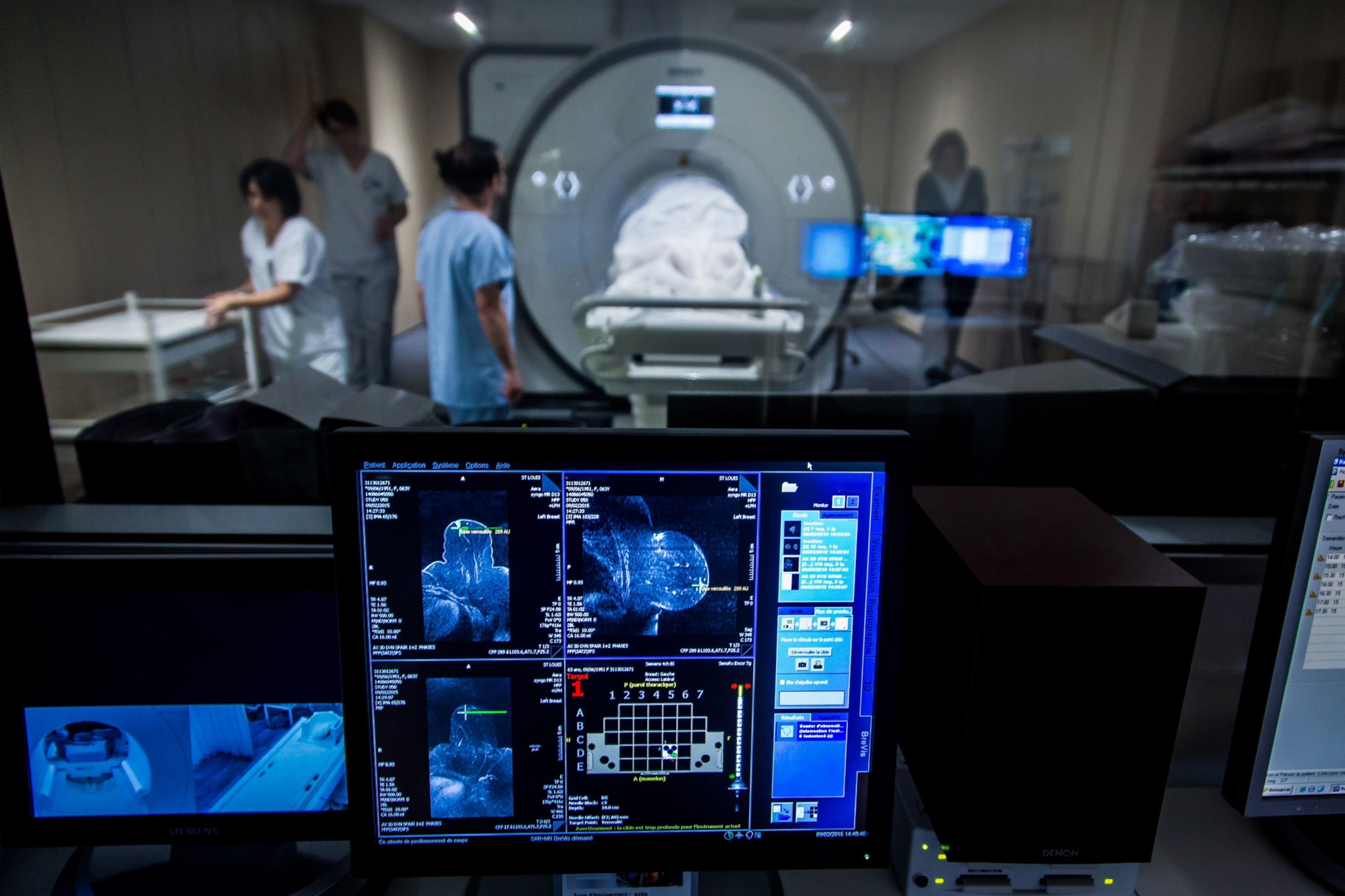 A computer monitor sits in the foreground, displaying blue MRI scans to guide surgical biopsy. In the background, partially out of focus, stands a large MRI machine with a patient and medical staff.