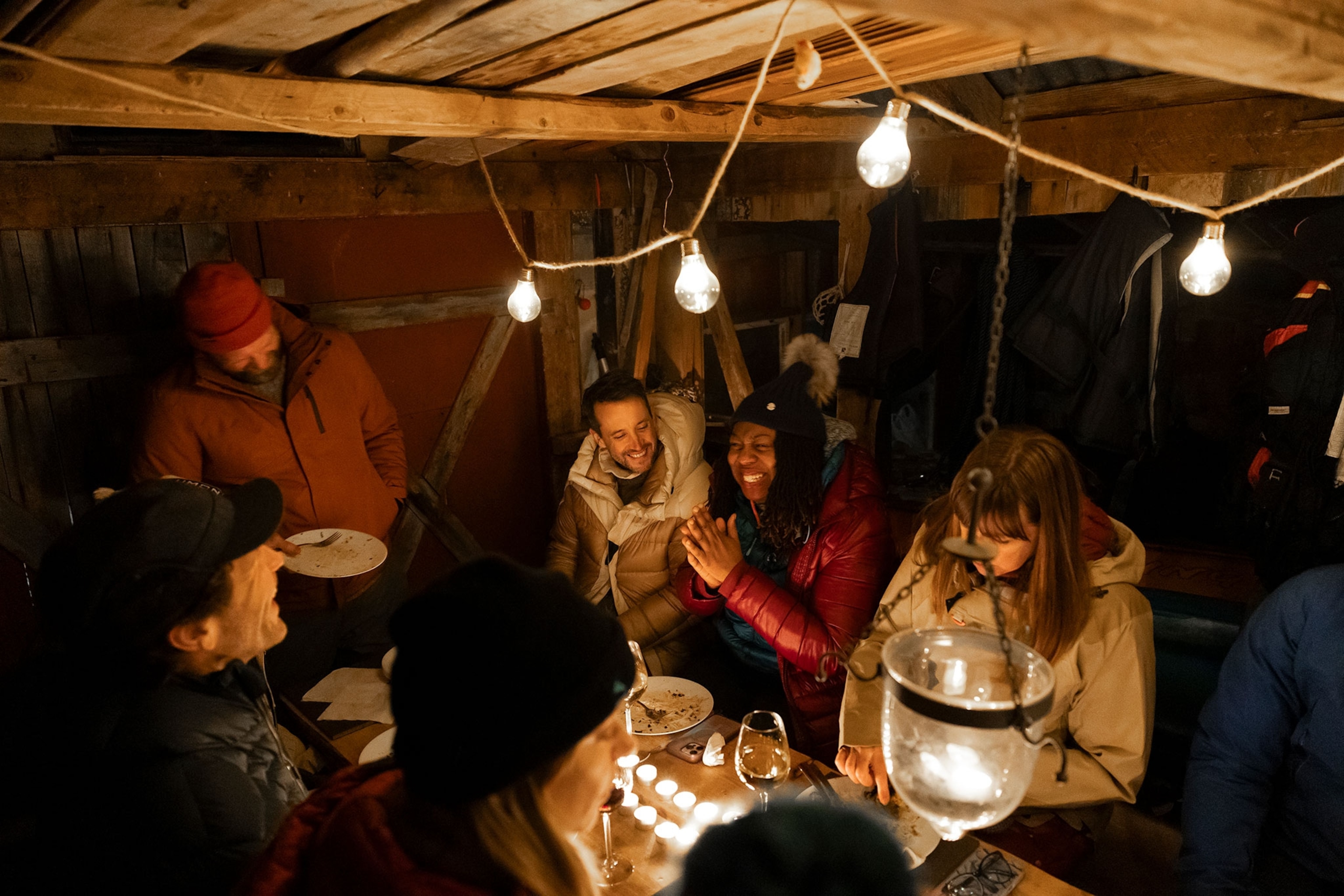 A group of people laugh at a dinner table in a dark cabin, lit by candles and string lights above.