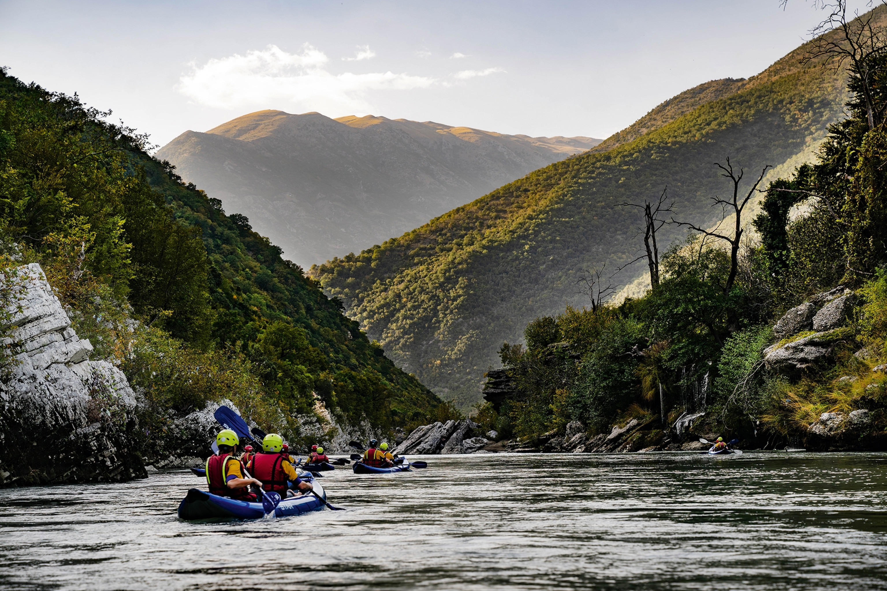 A group of rafters row upstream. The hills on either sides are coverage in plant matter, and a bare mountain can be seen in the far distance.