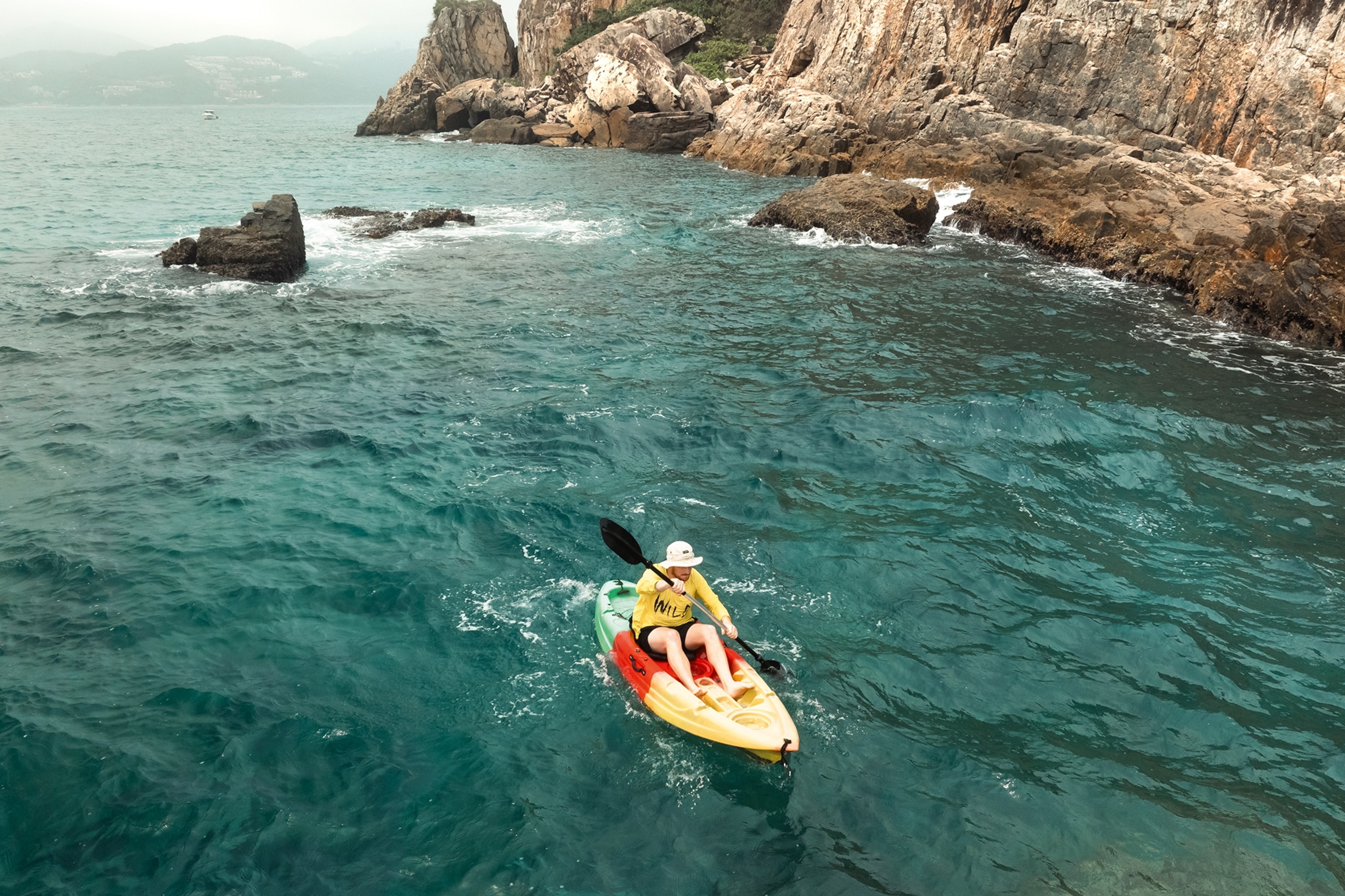 A lone kayaker in the clear waters off a cliff-hugged coastline.