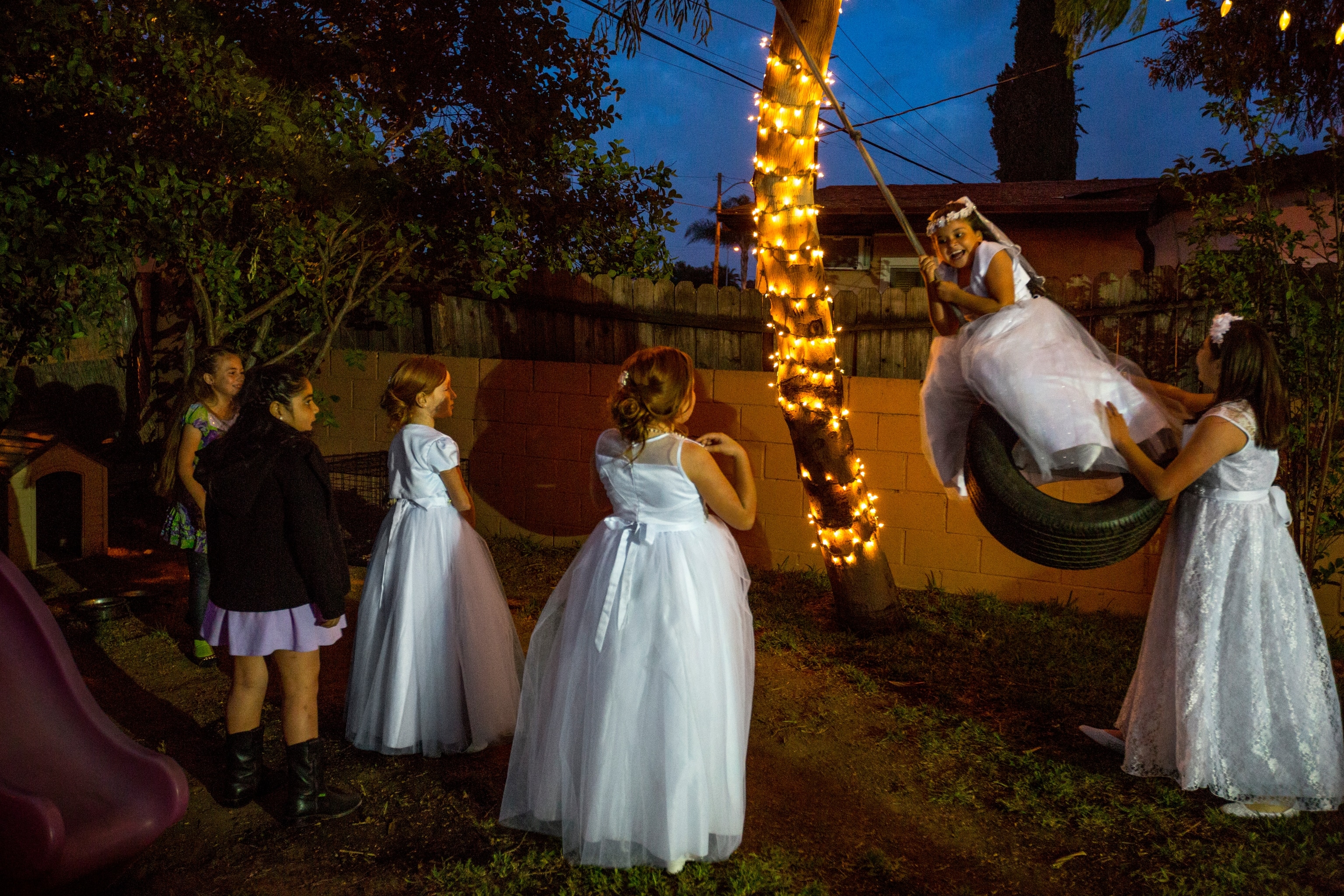 Six young Hispanic girls in communion dresses play outside at night, one happily being pushed on a tire swing as the others watch.