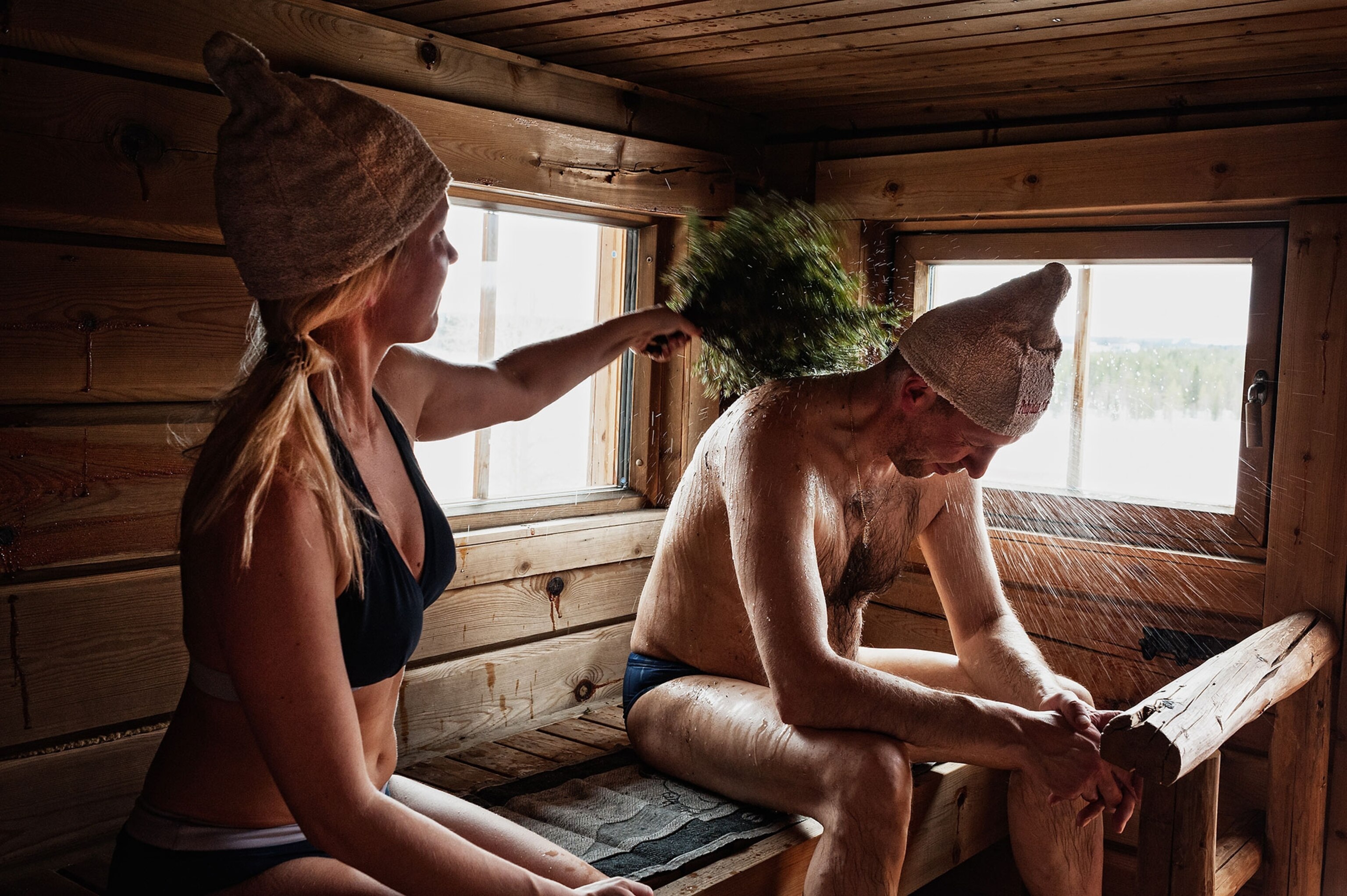 A woman in a bikini bathing suit hits a man wearing swim shorts with a bushel of leaves inside a wooden sauna