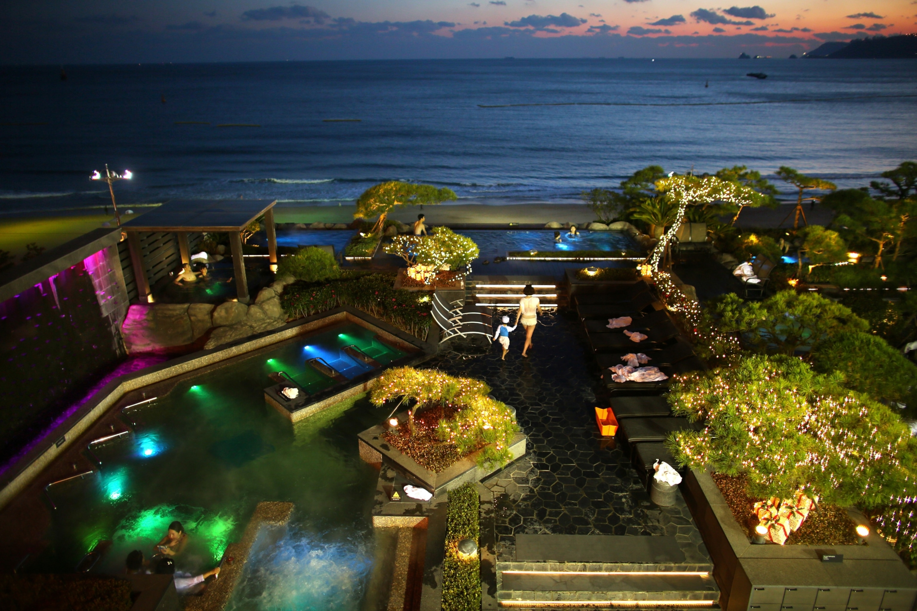 A view looking down at spa goers enjoying the various bathing pools overlooking a beach at sunset.