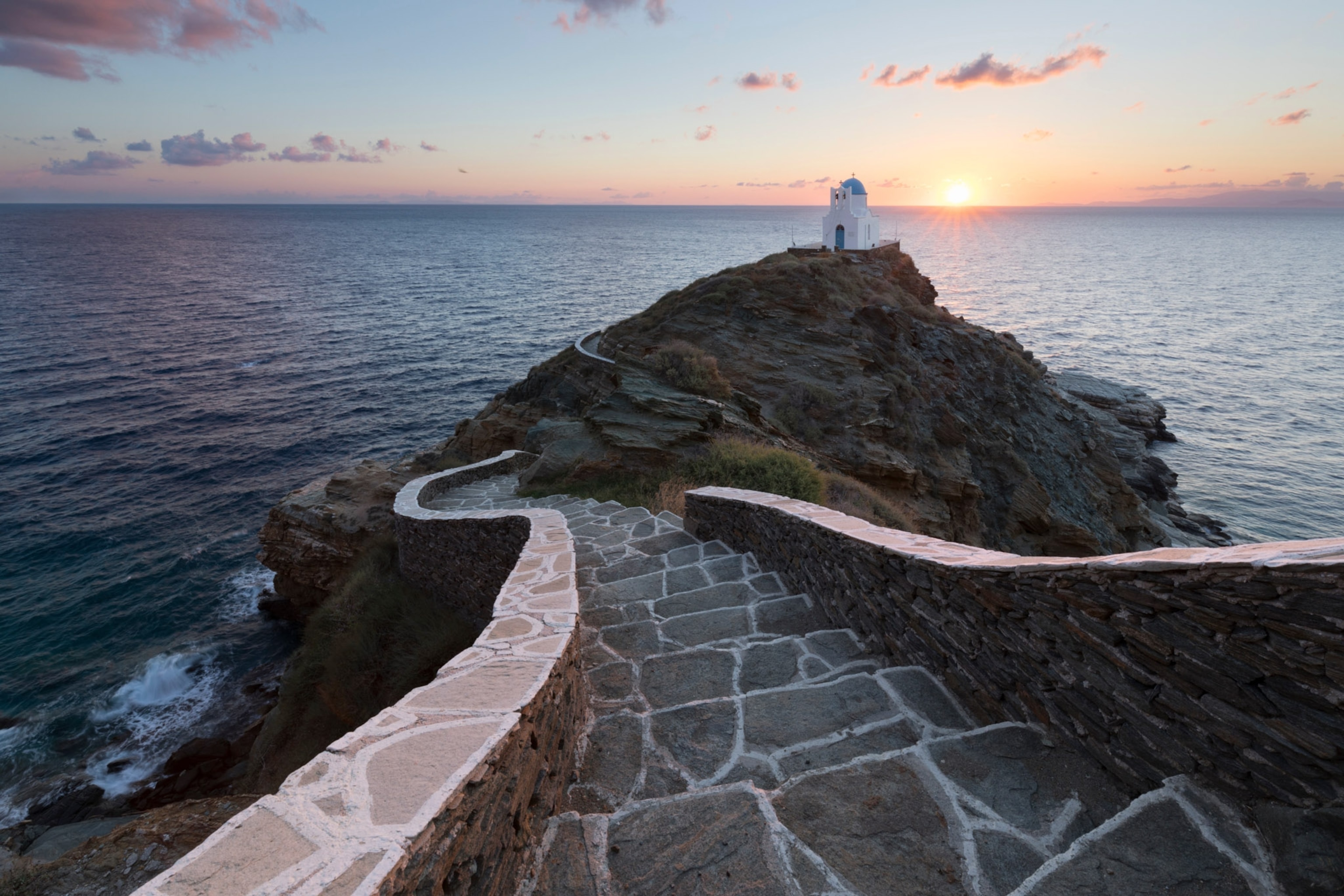 The photo stares down a pathway that with a short stone wall along each side and brush along the cliff. The sea and sunset sit behind the church.