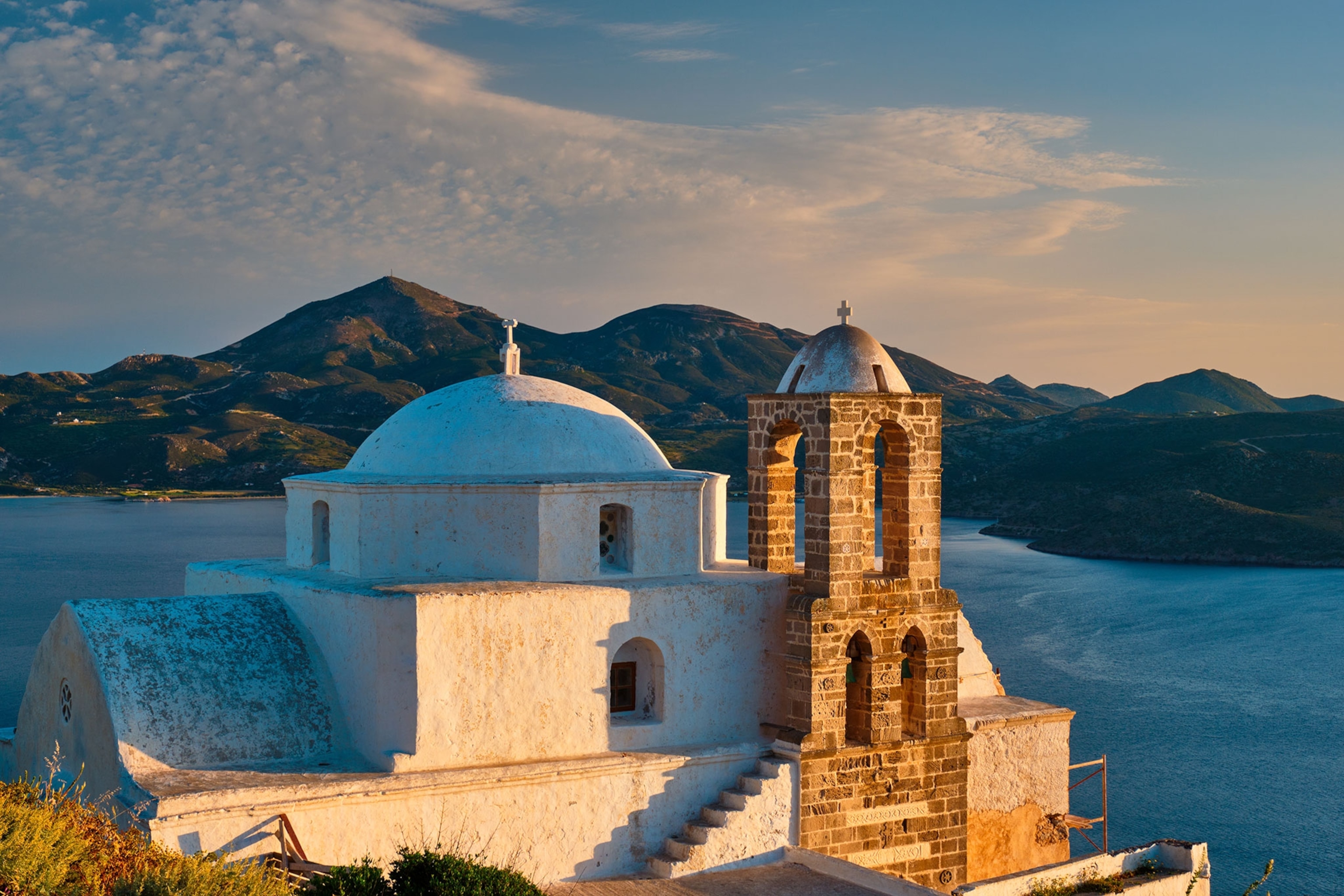 A white church sitting on a hilltop above water with mountains in the background during sunset.