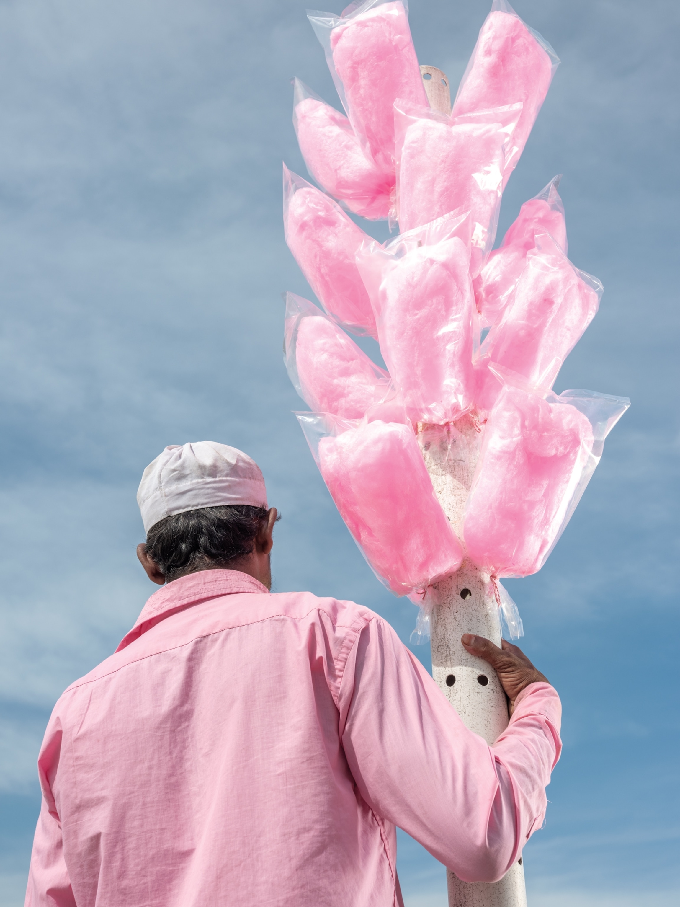 Picture of man in pink shirt seeing from his back, holding a pole with pink cotton candy in plastic wrap on display.