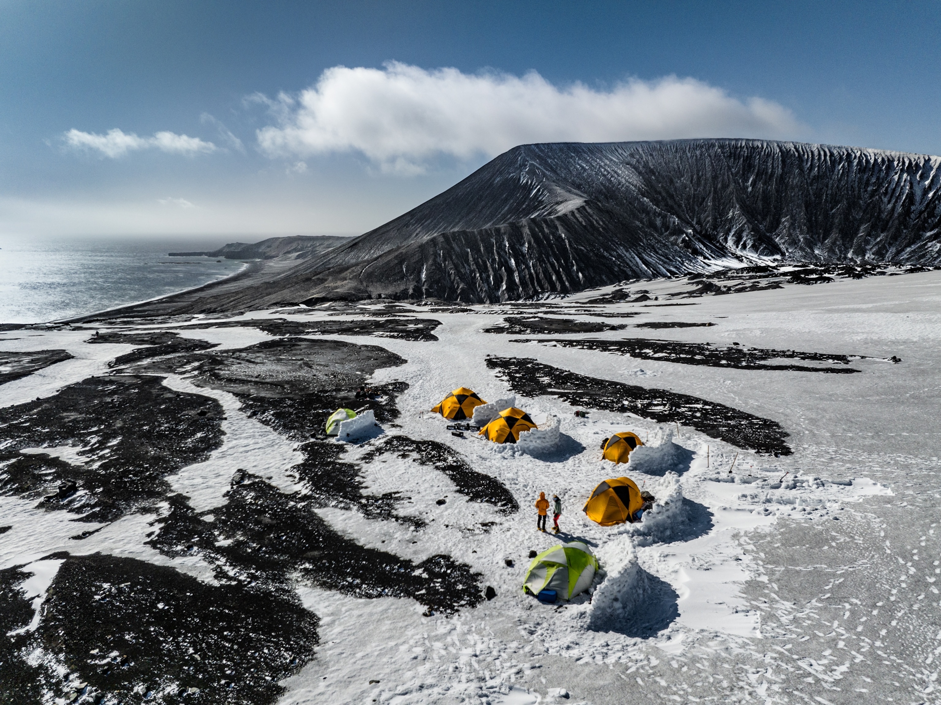 Drone shot of ashen hills with six bright colored tents protected from wind by walls built of blocks of snow and ice.