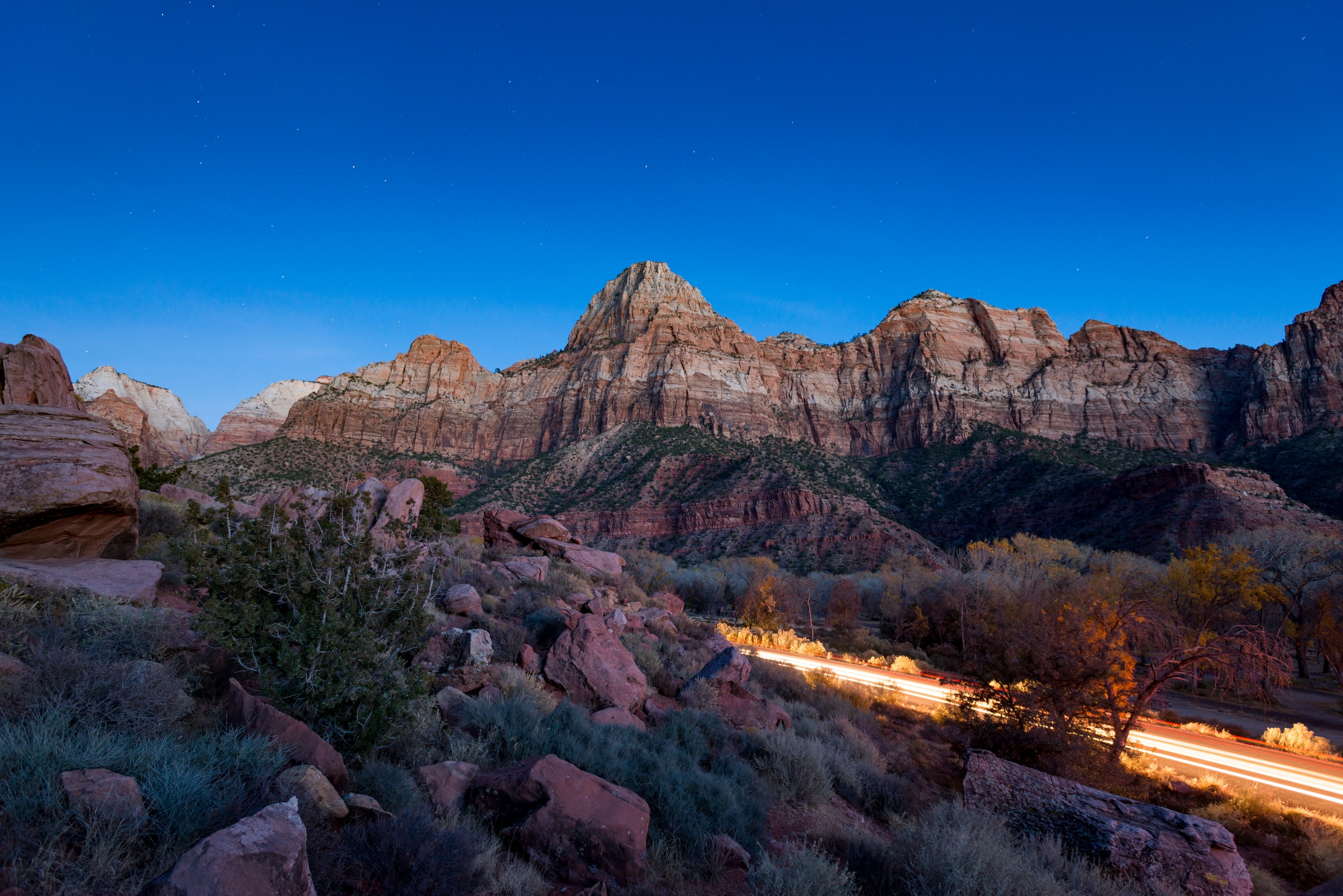 The steep red cliffs of Zion National Park glow in the moonlight as car headlights pass.