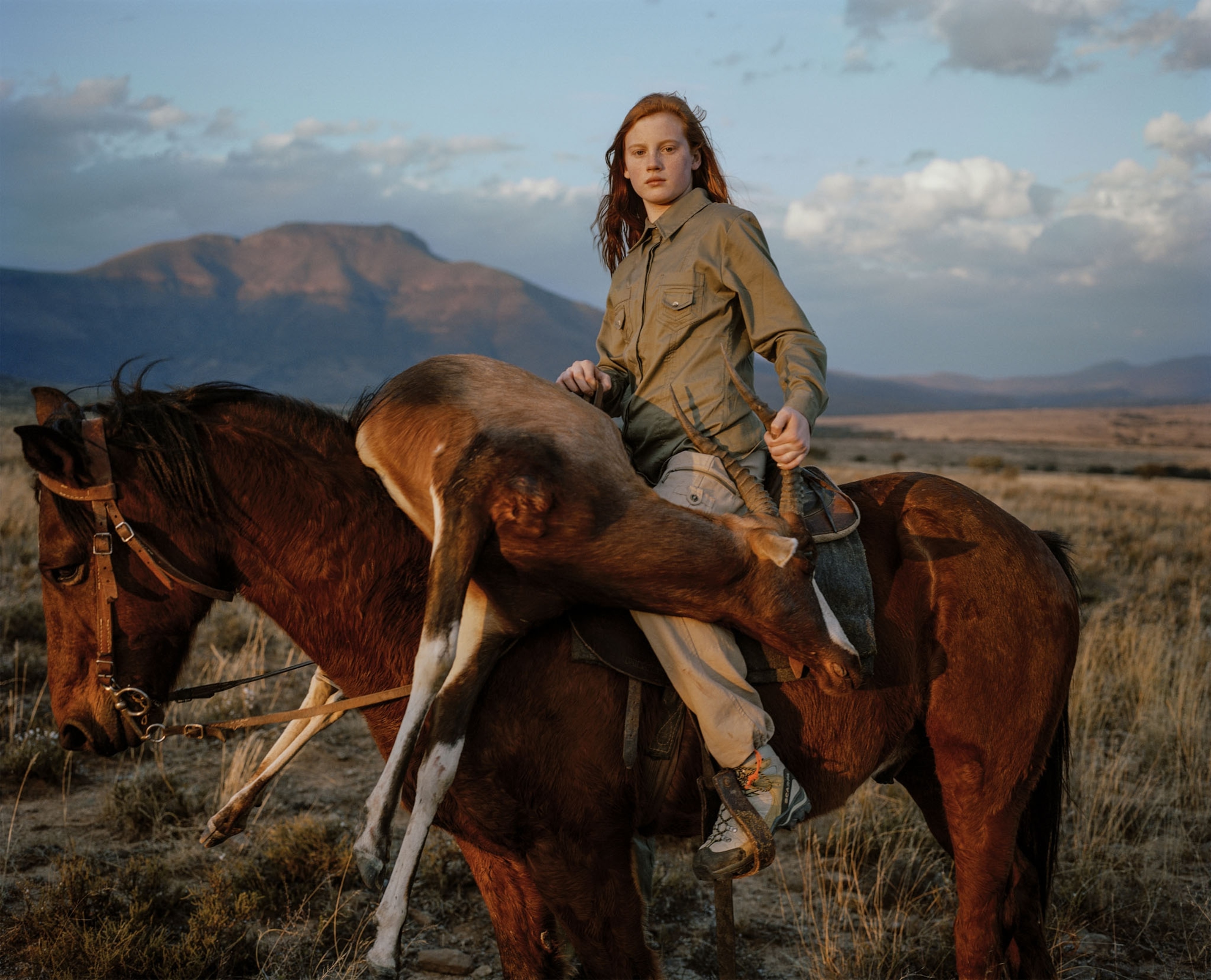 a red-headed girl on a horse with a dead horned animal strewn across the front of horse