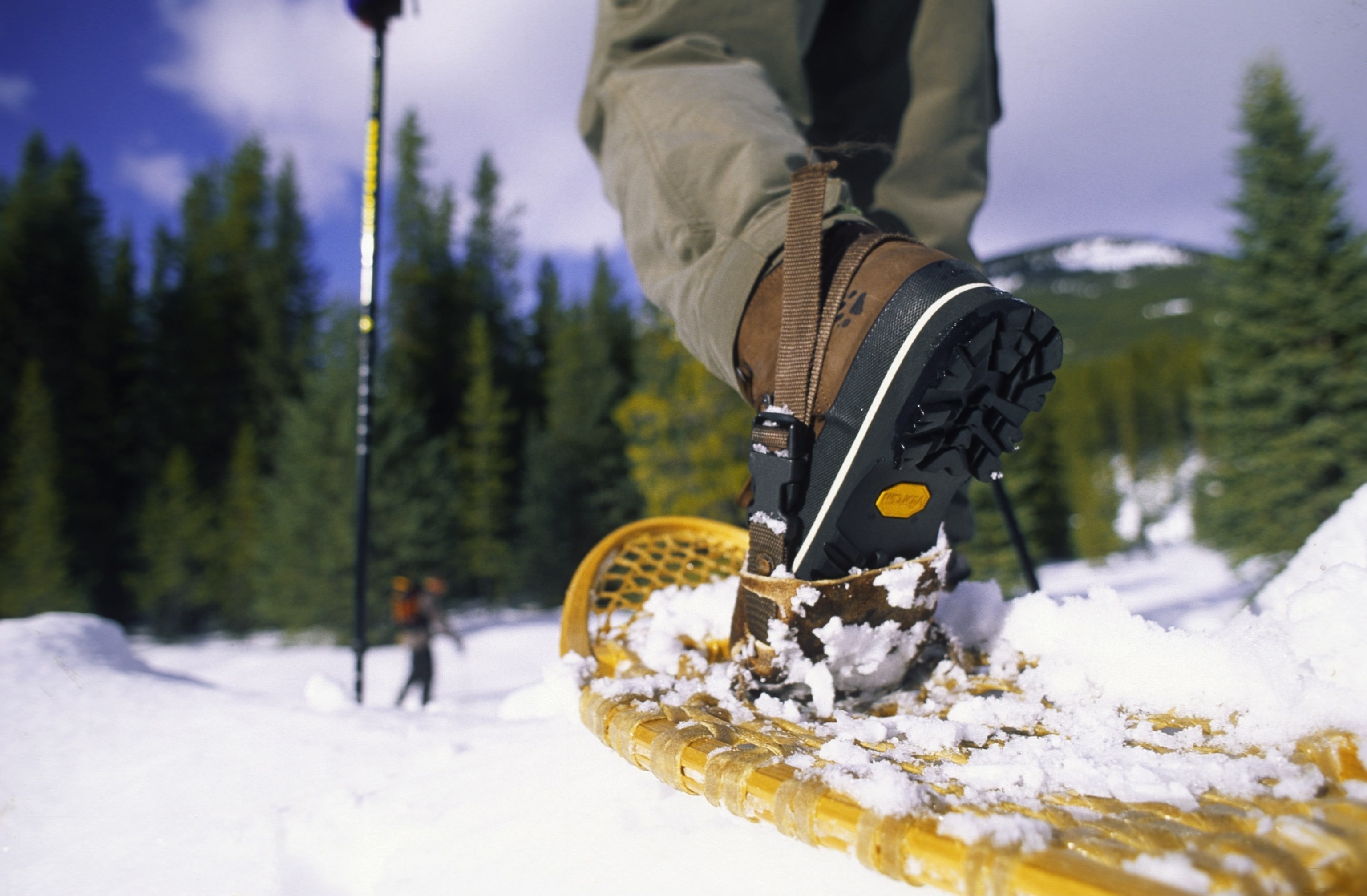 Boots strapped to a wooden snowshoe by the toe as a hikers walks through the forrest