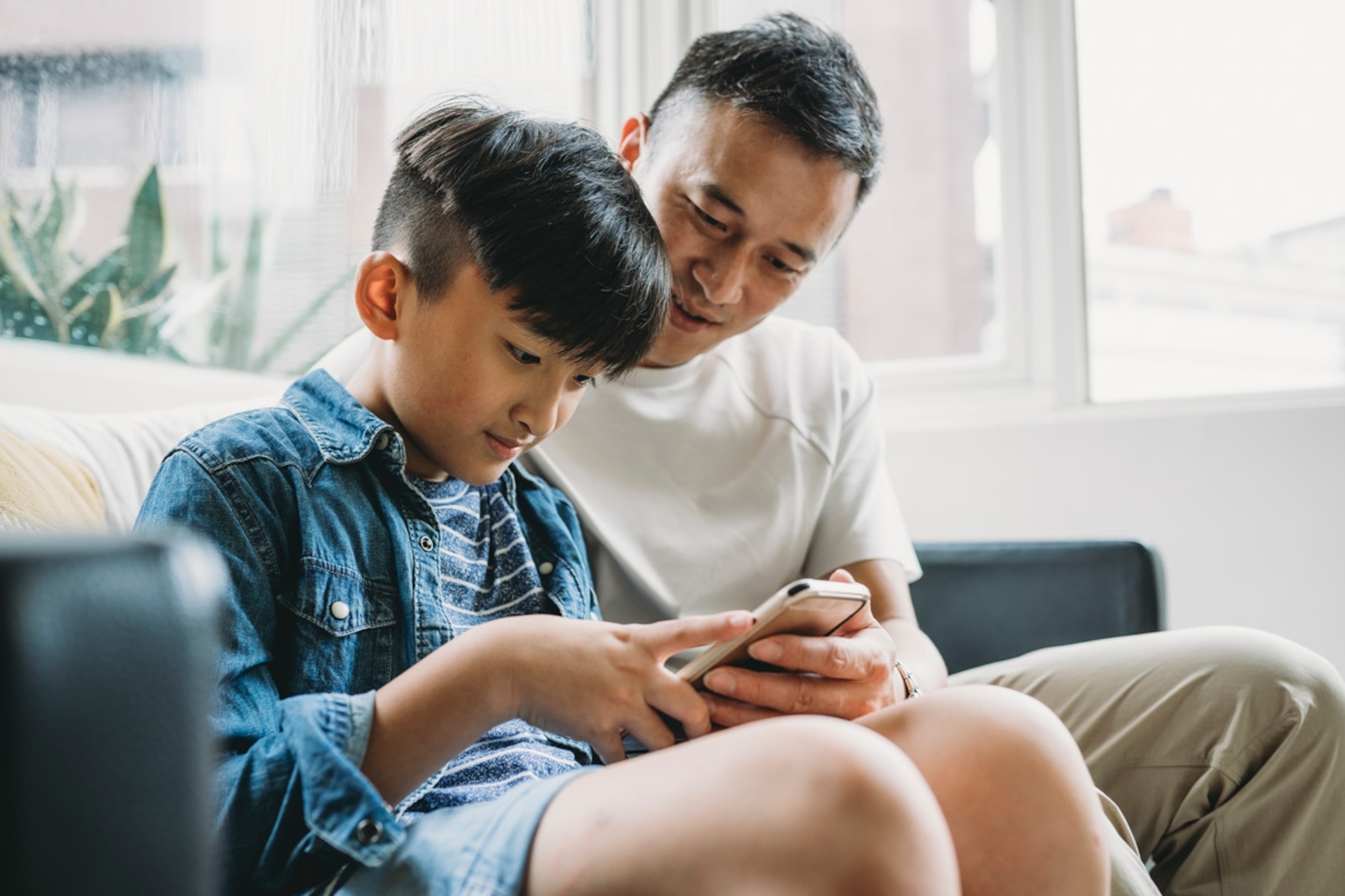 Father and son using a mobile phone together sitting on the sofa at home. Taiwan
