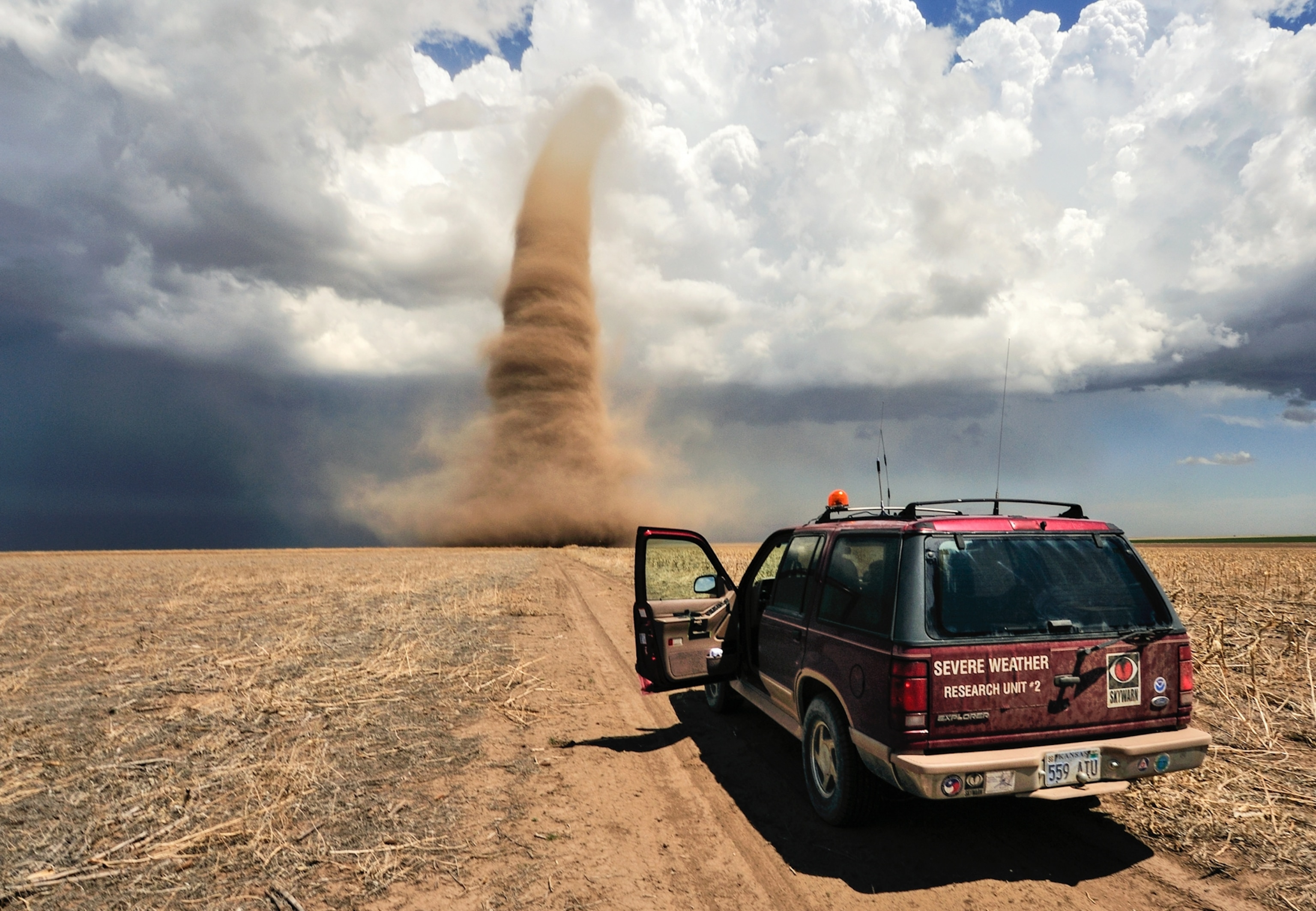 Professional storm chasers in a red SUV monitor an approaching tornado, which appears as an enormous swirling column of dust that disappears into the clouds.