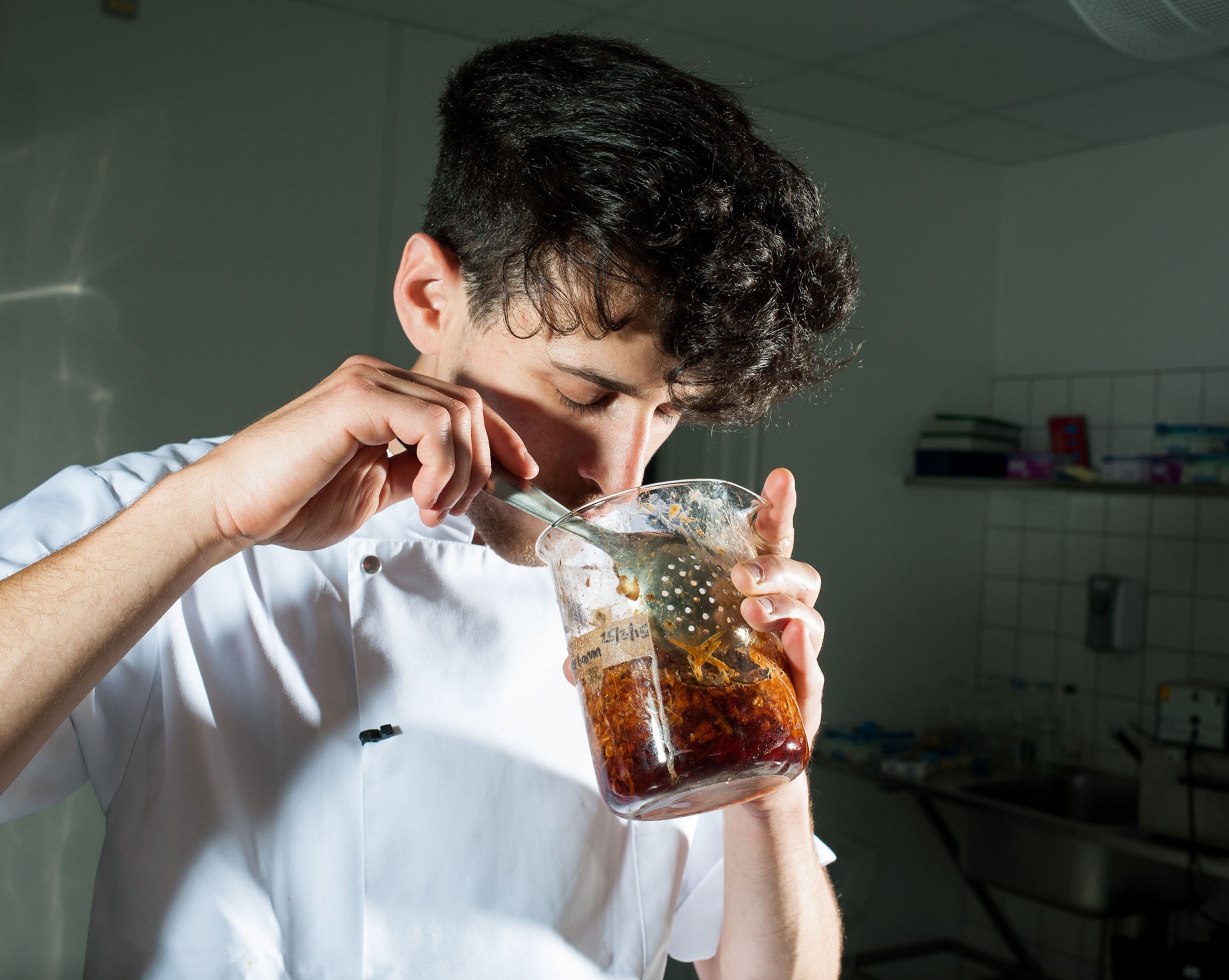 A man in a lab coat smells food in a large, round, glass beaker.