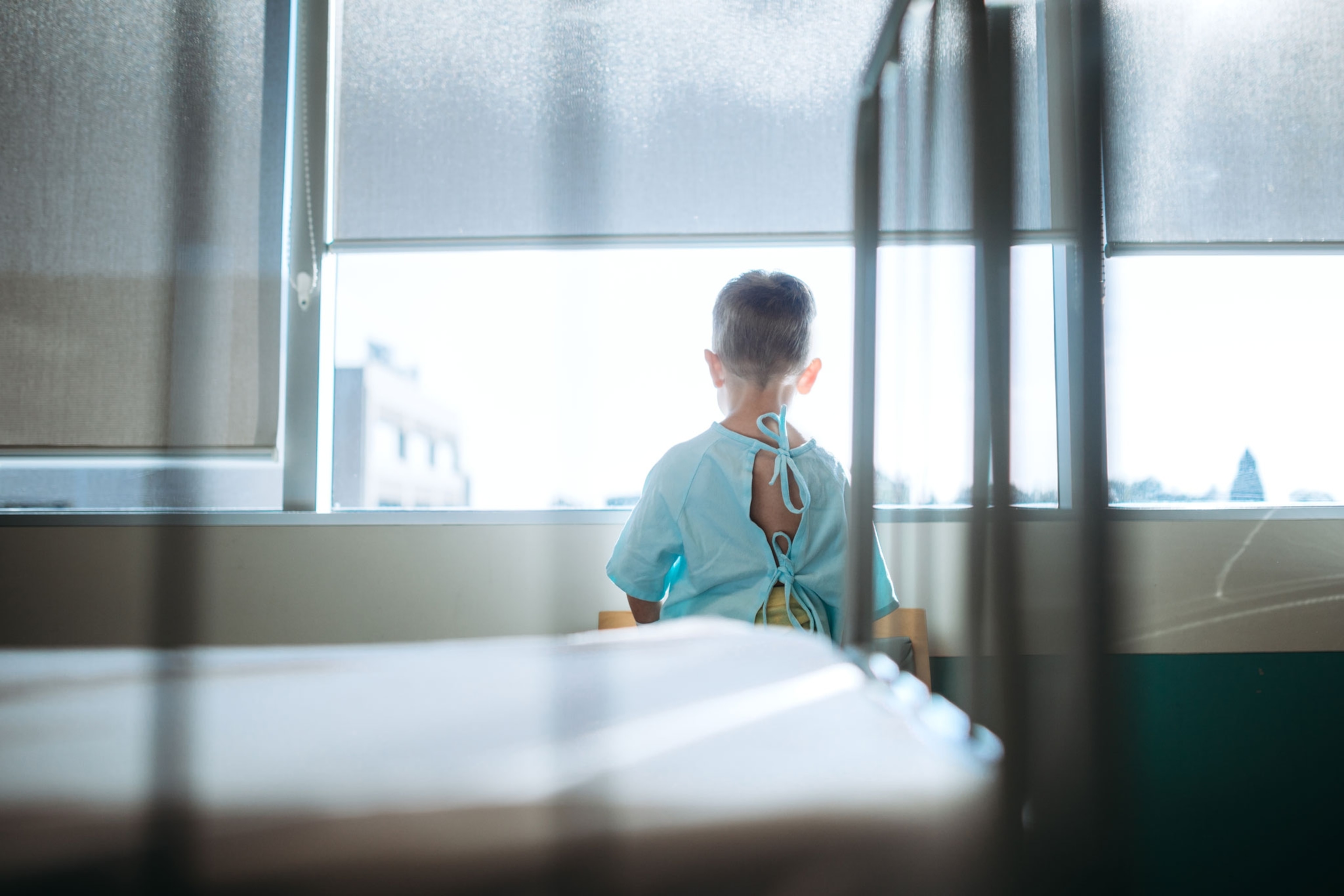 A cute little Caucasian boy waits in the childrens hospital ward to be taken back for surgery. He looks out of the window of his room while waiting for the doctor to arrive.