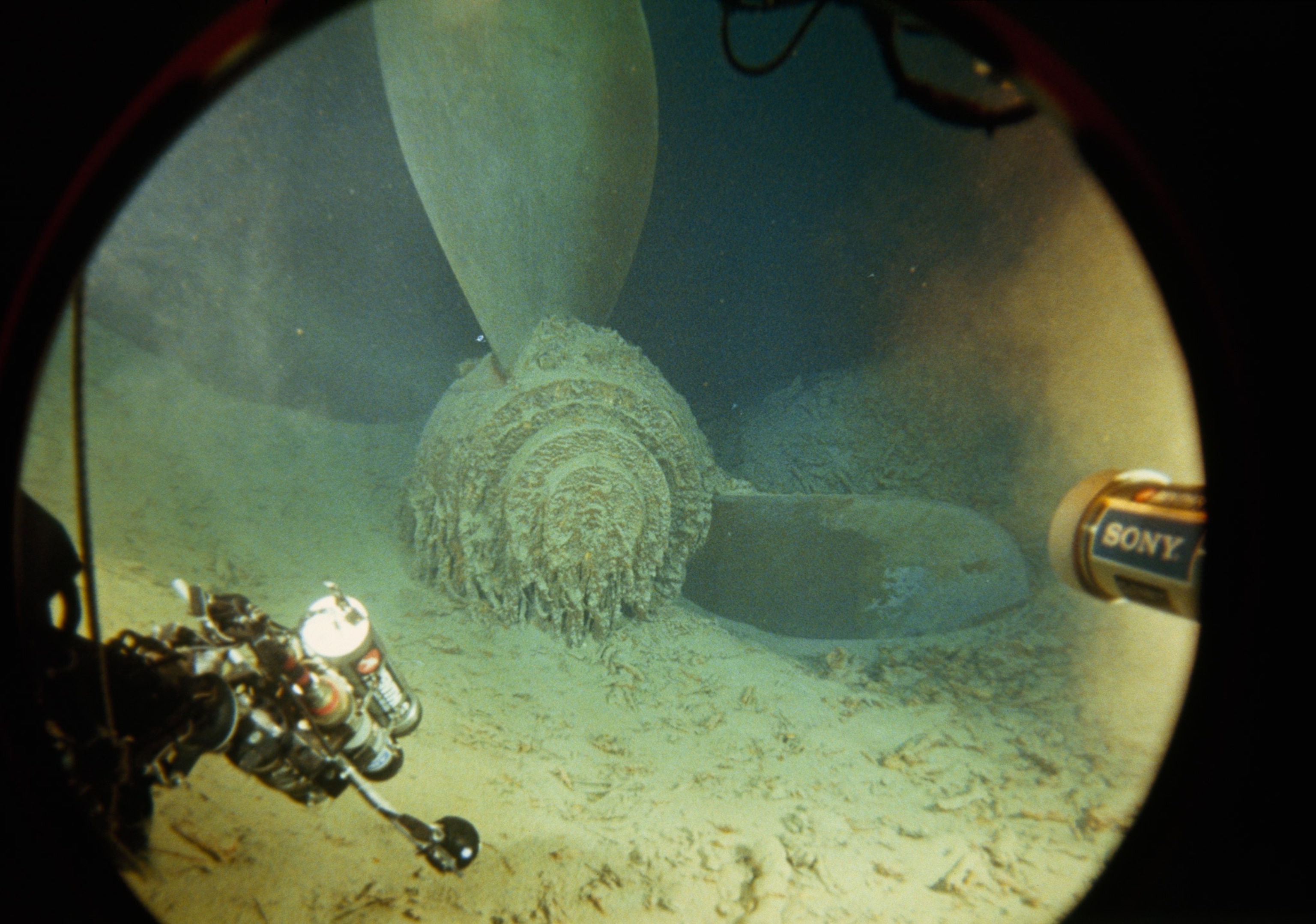 A light illuminates a propeller from the Titanic framed by the circular porthole of the submarine.