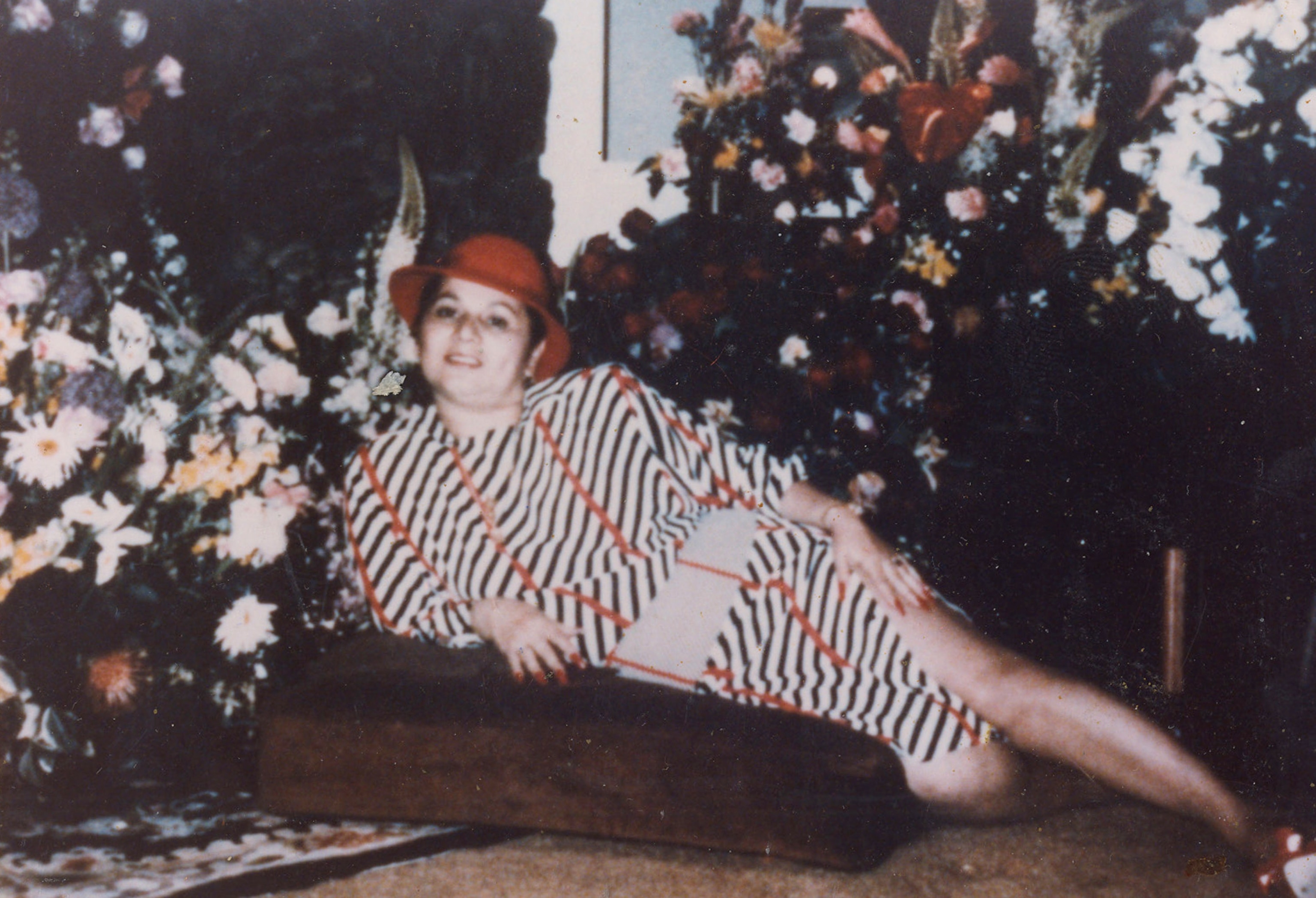 A womon in a dress patterend with overlaping stripes in red black and white lays on a cushion brown cushion cntered around a flowers that fill the background behind her.
