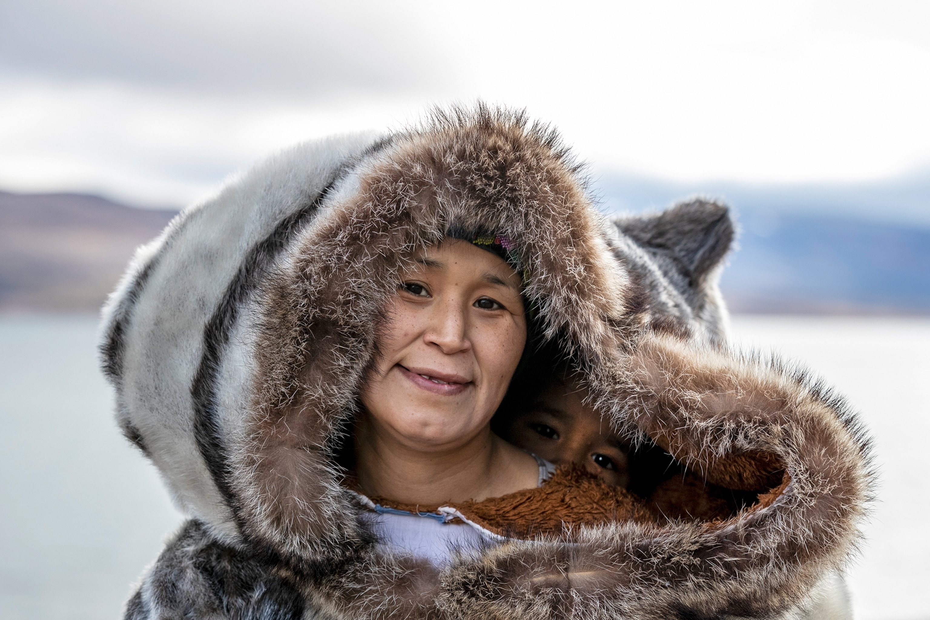 A woman is wearing a fur hooded parka, her child is visible inside the hood.