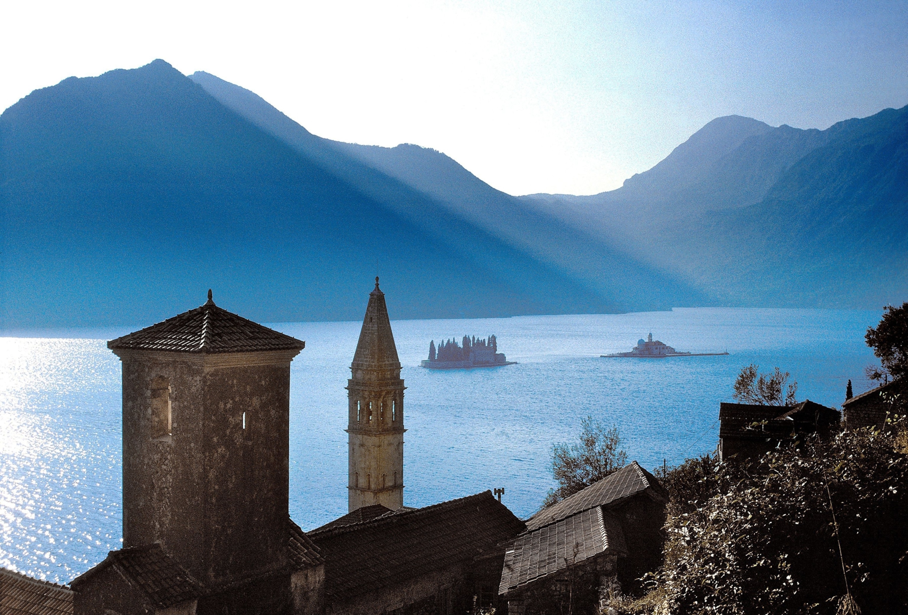 The islets of Sveti Dorde ans Gospa od Škrpjela are visible off the coast of Perast, as crepuscular rays shine down on the glistening waters.
