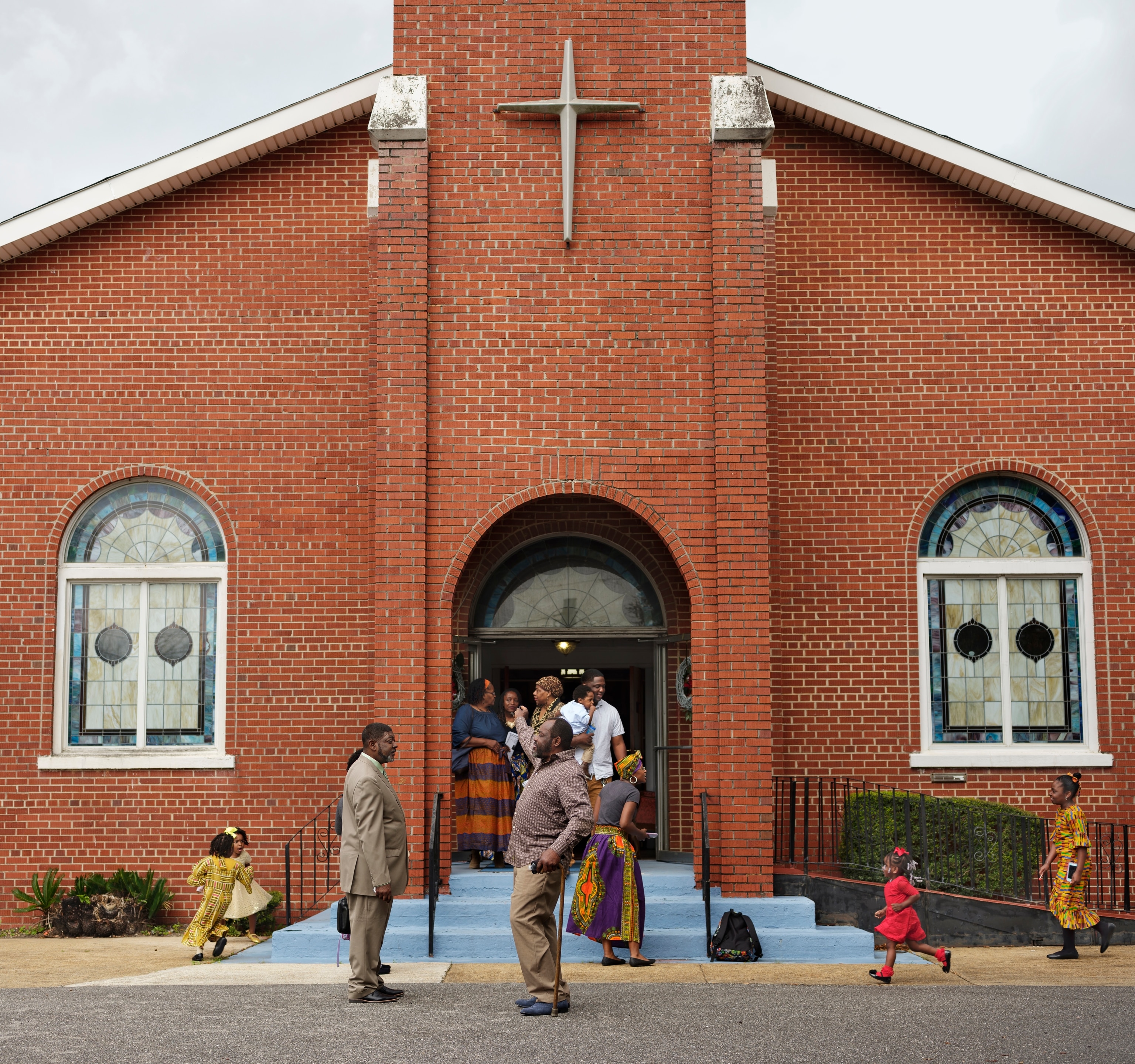 A small group of Black men, women, and children talk and play outside of a red brick church featuring a simple cross on its front.